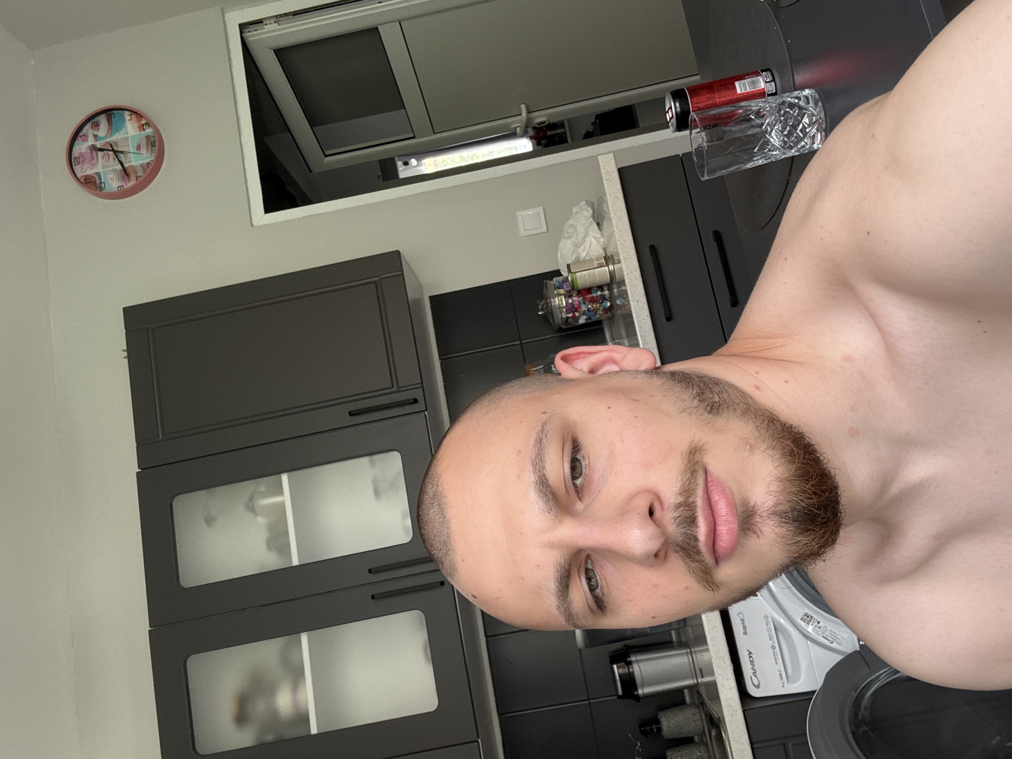 A shirtless man with a short buzz cut and a beard is taking a selfie in a kitchen. The kitchen has gray cabinets with frosted glass doors, a gray countertop, and a window. A red can and a glass are on the countertop near the man. A clock is on the wall. A washing machine is visible in the lower right corner. The location appears to be an apartment in Stara Zagora, Bulgaria.