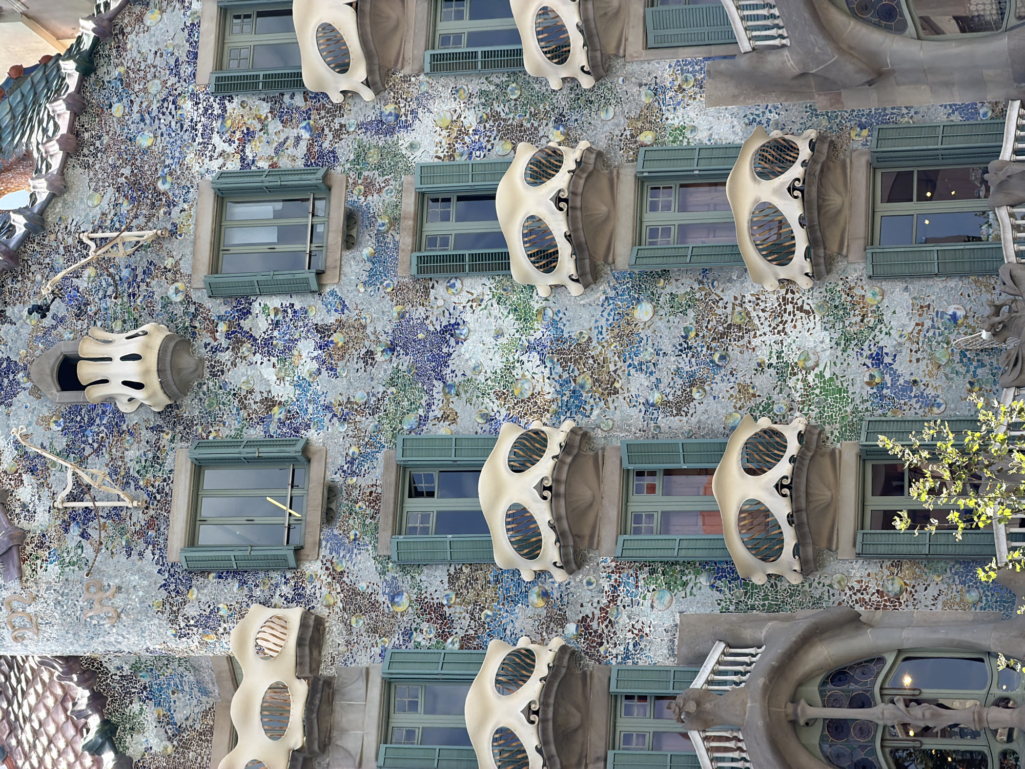 This is a photograph of the facade of Casa Batlló, a building in Barcelona, Spain, designed by Antoni Gaudí. The building is known for its organic, colorful, and whimsical architecture.
The facade is covered in a mosaic of broken ceramic tiles in various shades of blue, green, purple, and gold, creating a shimmering and iridescent effect. This mosaic is punctuated by a series of windows and balconies. The windows are rectangular and framed by pale, bone-like structures that resemble masks or skulls. These mask-like elements have two large, oval openings, each filled with horizontal slats that suggest ventilation or some kind of internal structure. The overall effect is surreal and theatrical.
The photograph is taken from a low angle, looking up at the building. The sunlight appears to be bright, casting shadows and highlighting the textures and colors of the facade. The presence of a leafy tree branch in the upper right corner indicates an outdoor setting and suggests it might be daytime. The architectural style itself evokes a sense of fantasy and playfulness. There is no visible text in the image. The building's unique design suggests it is a tourist attraction or a significant architectural landmark.