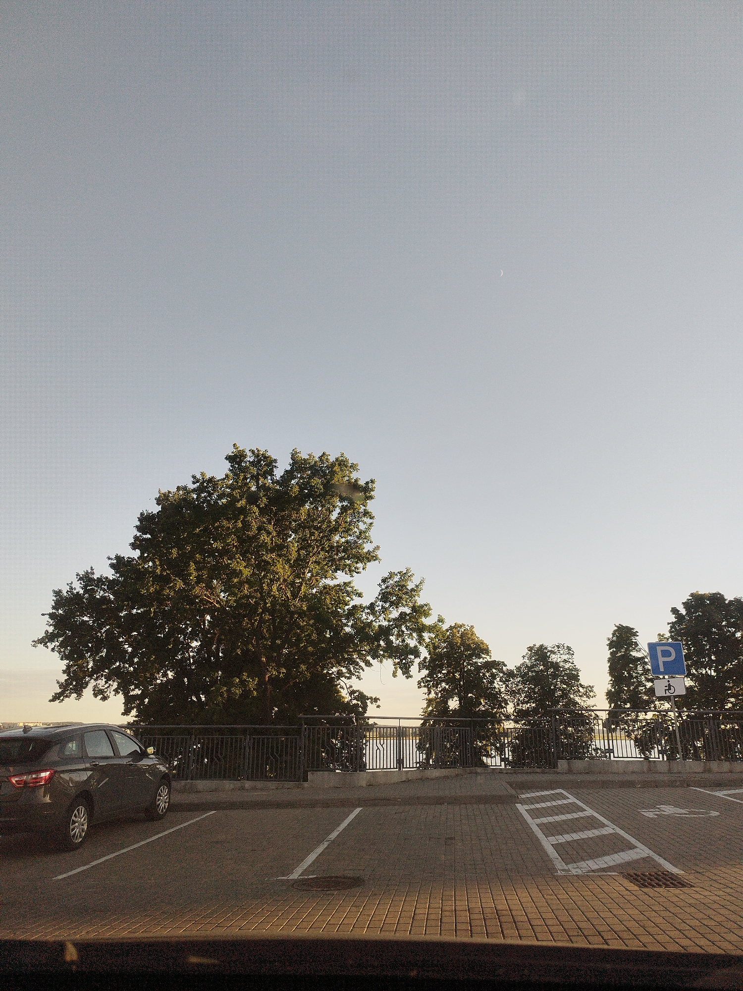 An eye-level, exterior shot shows a parking lot scene with a clear sky and a line of trees. In the foreground, a dark-colored car is parked in a designated spot marked with white lines. The parking lot pavement is made of interlocking bricks and asphalt. A metal fence separates the parking lot from a walkway or embankment, beyond which several large trees are visible. A blue parking sign with a white "P" and a wheelchair symbol is mounted on a pole in the background, near the trees. A faint crescent moon is visible in the upper portion of the sky. The lighting suggests it is either early morning or late afternoon.