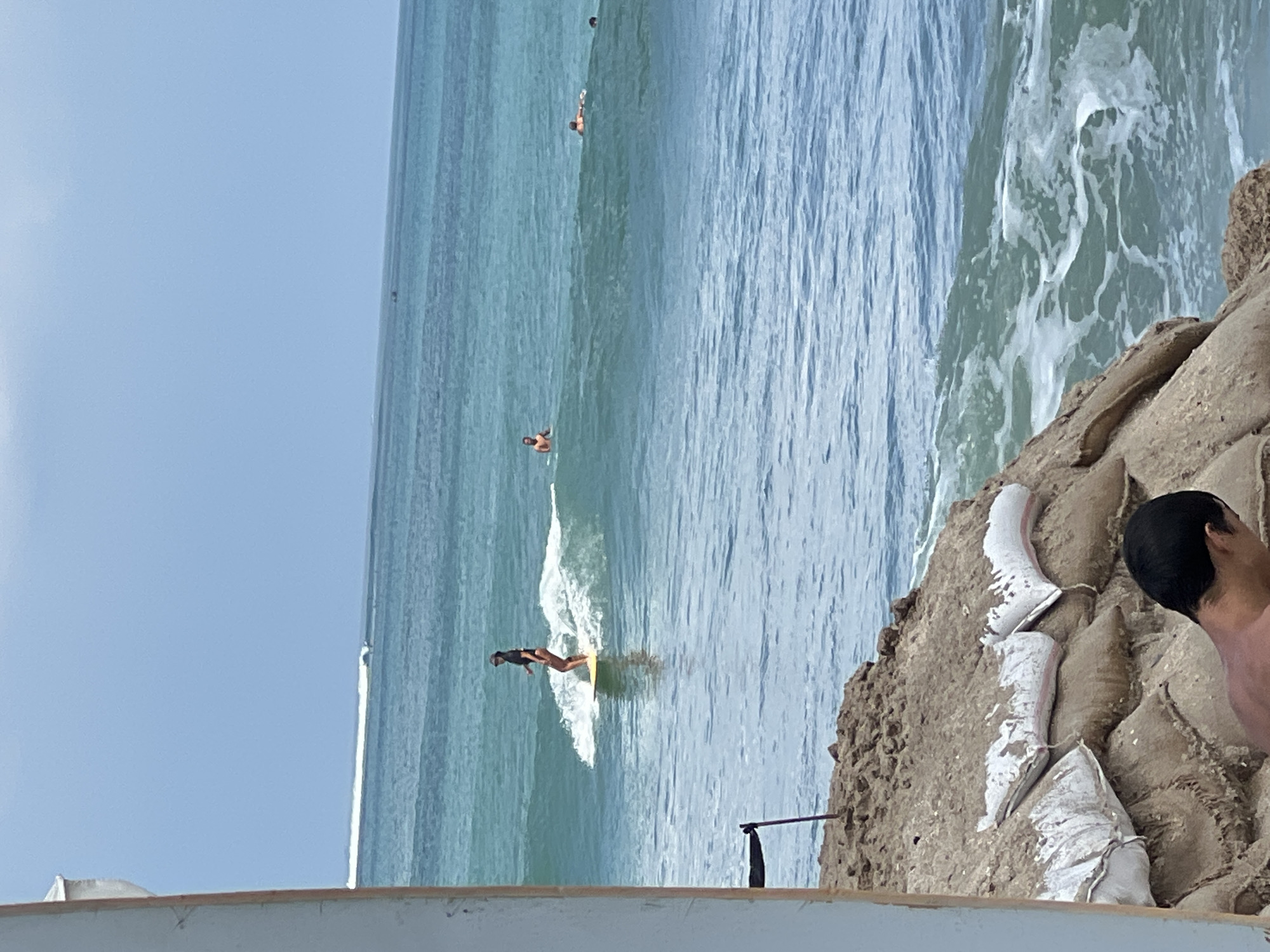 The image depicts a beach scene in Kuta, Indonesia, with several individuals engaged in surfing and swimming. In the foreground, a person's back is visible, facing away from the camera and towards the ocean. The middle ground shows a woman actively surfing on a yellow board, riding a wave with white foam. Further out in the water, three other individuals are present; one is standing in the water, another appears to be lying on a surfboard, and a third is partially submerged. The ocean is a vibrant turquoise, with visible waves and ripples. The background is dominated by a clear blue sky. On the shore, a rocky embankment is reinforced with sandbags, some of which are covered in white material, suggesting a protective barrier against erosion. The overall impression is a sunny day at a popular surf spot.