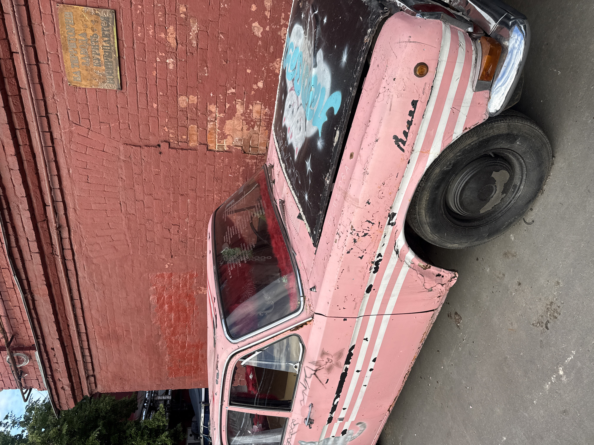 A faded pink car with peeling paint and white stripe decals is parked on a paved surface in front of a weathered, red brick building in Moscow, Russia. The car's hood is painted black and features a spray-painted image of two white cats with blue accents and small white stars. The car's windows are tinted pink, and the interior appears to be decorated with pink fur. A yellow sign with Cyrillic text is affixed to the brick wall. The sky is partly cloudy.