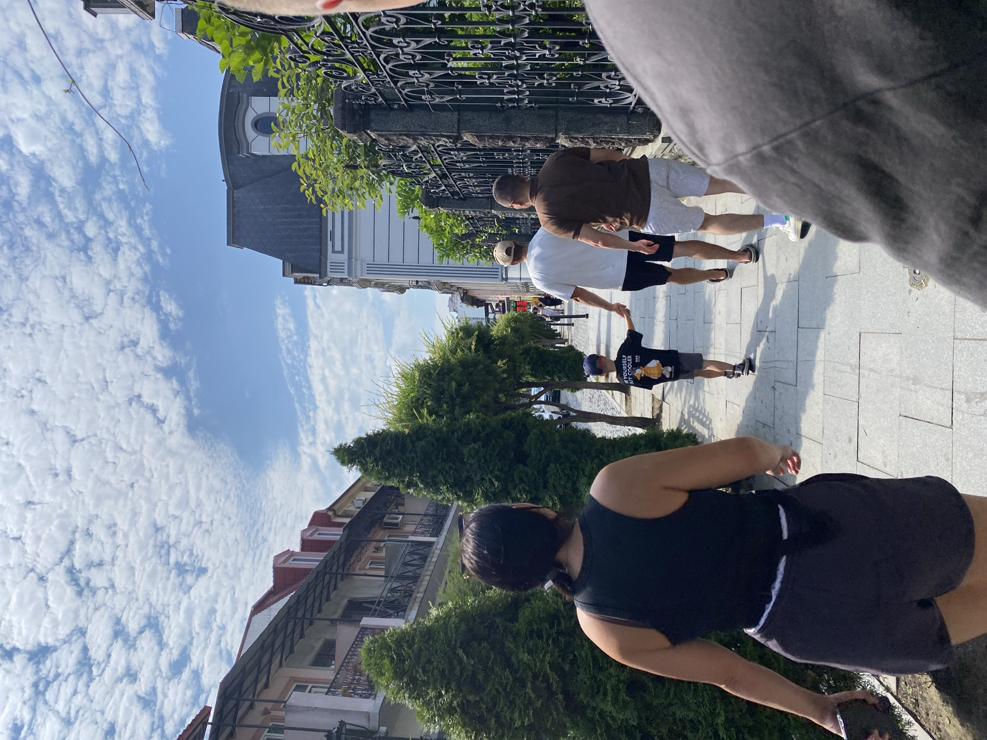 A group of people are walking down a street in Batumi, Georgia. In the foreground, a woman with dark hair wears a black tank top and gray shorts. She is walking away from the camera, holding a dark-colored object in her left hand. Behind her, a young boy in a black t-shirt and gray shorts walks holding hands with a man in a white t-shirt and black shorts. To their right, another man wears a brown t-shirt and light gray shorts. They are walking on a paved sidewalk lined with trees and buildings. The buildings on the left appear to be older, with balconies and red-tiled roofs. On the right, a decorative black metal fence encloses a green area with more buildings in the background. The sky is blue with scattered white clouds. The scene suggests a sunny day in a pedestrian-friendly area of the city.