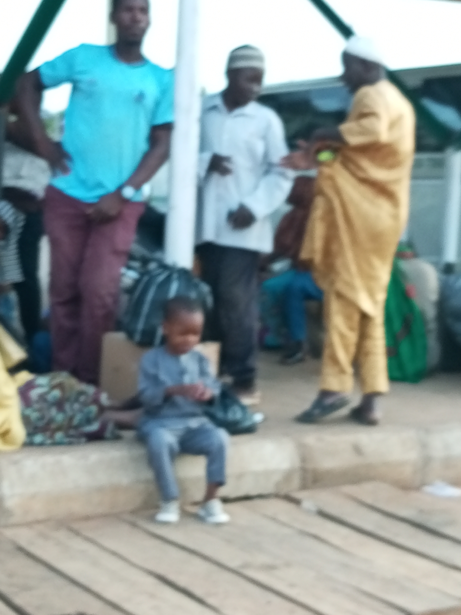 In Gombe, Nigeria, a group of people are gathered on a paved surface with wooden planks in the foreground. In the center, a young child, appearing to be a boy, is sitting on a raised concrete ledge. He is wearing a grey, long-sleeved outfit and white sneakers, and is looking down at something in his hands. To his left, a man stands with his hands on his hips, wearing a bright blue t-shirt and maroon trousers. He has a watch on his left wrist. Behind him, another man is visible, wearing a white shirt and a patterned cap. To the right of the child, a person in a yellow robe and white cap is interacting with someone out of frame, holding a small, round object in their hand. Other individuals and bags are present in the background, with a green structure visible above. The lighting suggests it is daytime.
