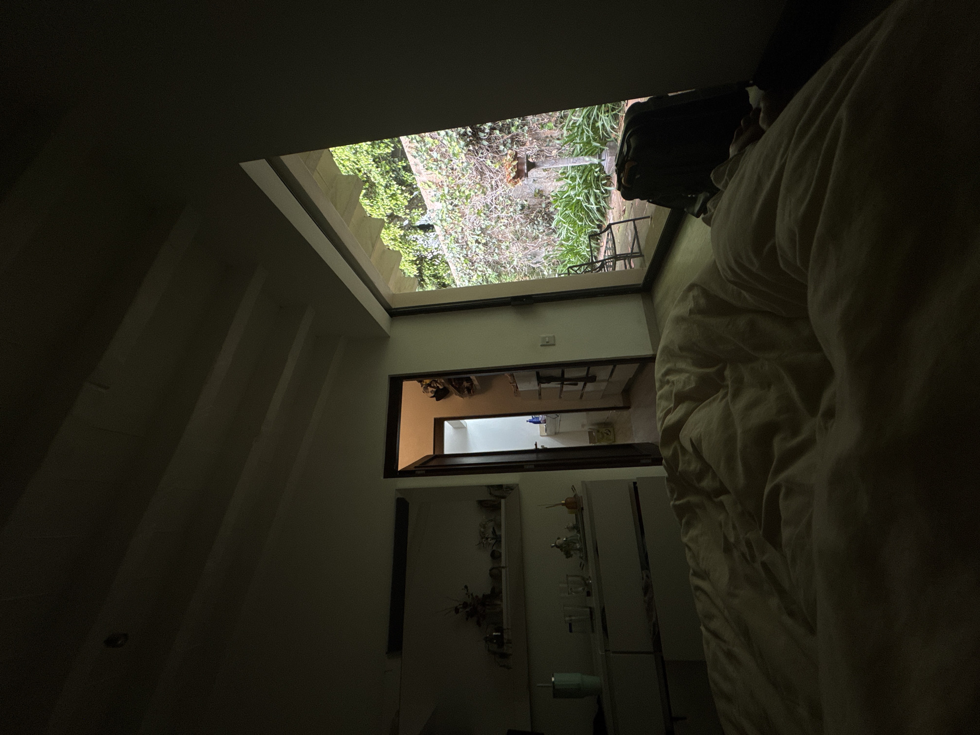 The photograph depicts an interior space, likely a room, leading through a series of openings to an exterior garden area. The foreground is dark, suggesting a dimly lit or shadowed portion of the room with a bed partially visible on the right side. A sequence of doorways or openings lead to increasingly brighter areas. The final opening showcases a garden with foliage and some structures. Based on the architectural style and vegetation type, it is potentially located in Mexico City, Mexico, but the exact location is not discernible from the image alone.