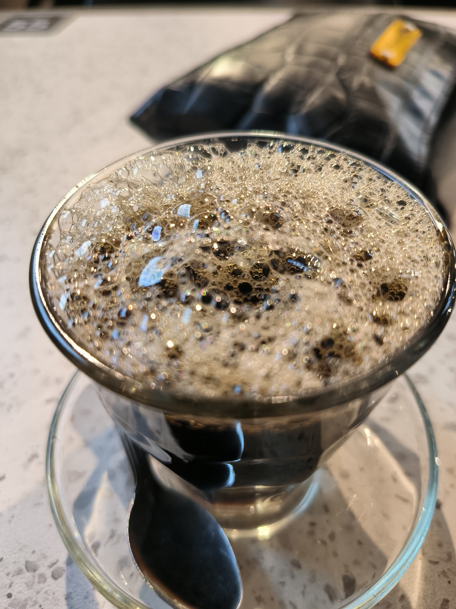 A close-up, overhead shot shows a glass cup filled with frothy coffee and a spoon on a saucer. In the background, a dark, crinkled object with a yellow tag rests on a speckled countertop. The image appears to have been taken in an indoor setting, possibly a cafe or kitchen. The location of Lhokseumawe, Indonesia is not depicted in this image.