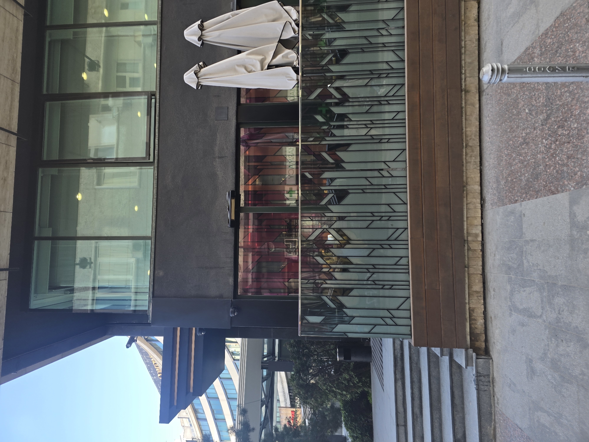 An outdoor scene in an urban setting, featuring a modern building facade with ground-level pavement. The building has a dark, likely black or dark grey, exterior. Its upper section features large, rectangular, reflective windows that show reflections of another building and internal light sources. Below these windows, a section of the building incorporates a glass railing with a distinctive geometric pattern of dark lines. Behind this glass, the interior of a pharmacy is reflected, visible through a green cross symbol and mirrored text "YCARMНАЧ" (PHARMACY).
In front of the building, there is an outdoor patio area constructed from wooden planks. Two light-colored, closed outdoor umbrellas with dark trim are positioned above this patio. The ground in the foreground is composed of different materials: a large section of light grey concrete paving, a reddish-brown speckled granite area, and concrete steps descending on the left side of the frame. A metal bollard or post is visible on the granite pavement.
The sky is bright and clear blue, suggesting daytime. Reflections in the glass surfaces and visible elements in the background indicate other contemporary buildings and some greenery. The word "GOOSE" is embossed on the metal bollard. No people or direct activities are visible, presenting a still urban snapshot. The provided context places the location in Sofia, Bulgaria.