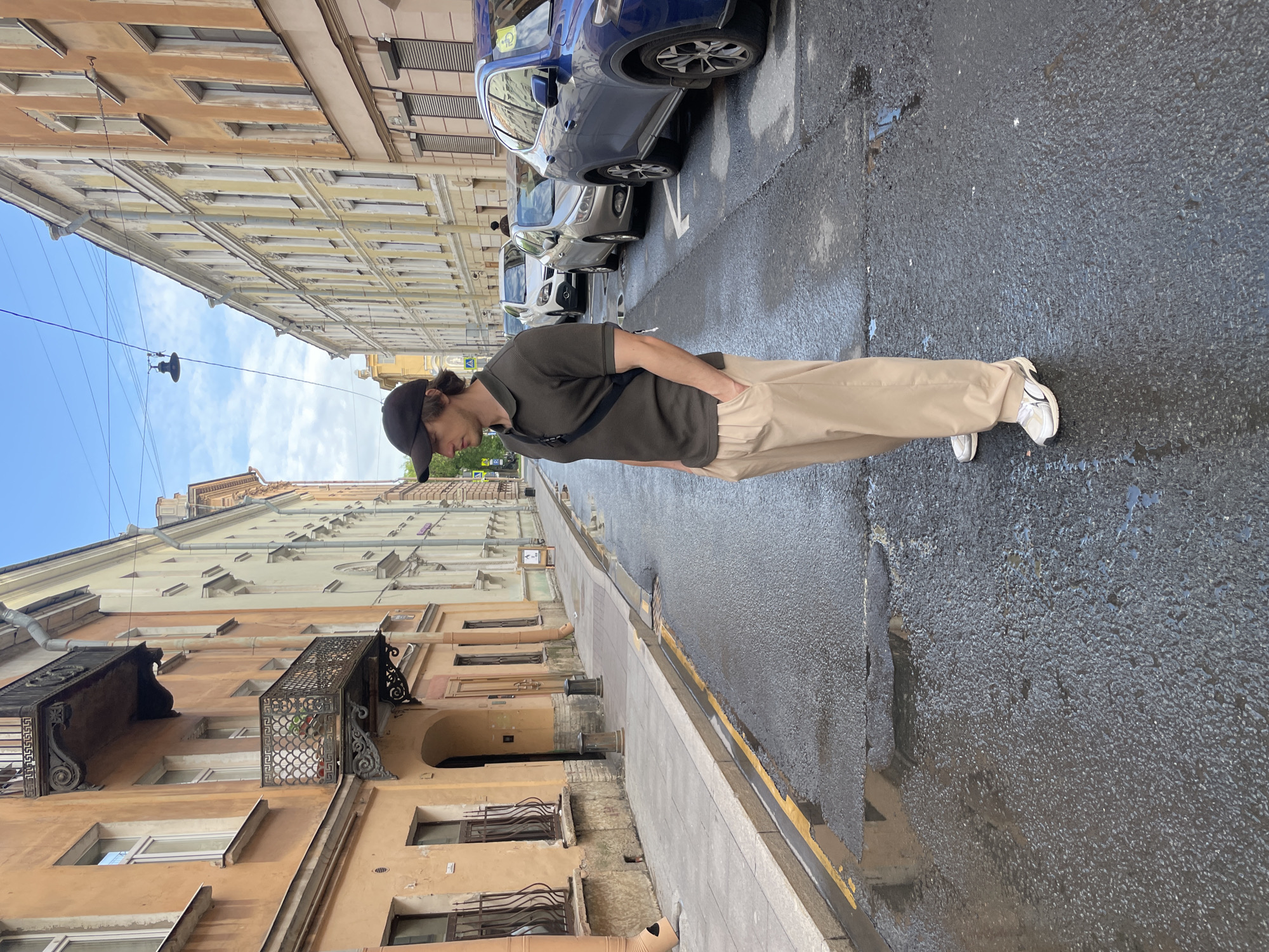 A full-shot, slightly low-angle photograph captures a young man standing on a wet urban street, consistent with the architecture found in Saint Petersburg. The man is positioned on the left side of the frame, facing away from the camera and looking slightly downwards. He wears a dark olive green polo shirt, light beige trousers, and a black baseball cap, with a black sling bag across his chest. His hands are casually tucked into his pockets.
The street appears to have recently experienced rain, as the asphalt road surface is visibly wet with some puddles and reflections. A white arrow is painted on the road, indicating a forward direction, and a yellow line marks the edge of the curb. To the man's right, several cars are parked along the street, including a blue SUV and a silver sedan.
Both sides of the street are lined with multi-story buildings, characteristic of historical European cities. These buildings feature ornate details, balconies with intricate wrought iron railings, and varied facades in shades of yellow, orange, and beige. Overhead wires traverse the street above, and a street light fixture hangs centrally. The sky is partly cloudy, indicating daytime. No legible text is visible, although small signs and a car sticker are present in the background.