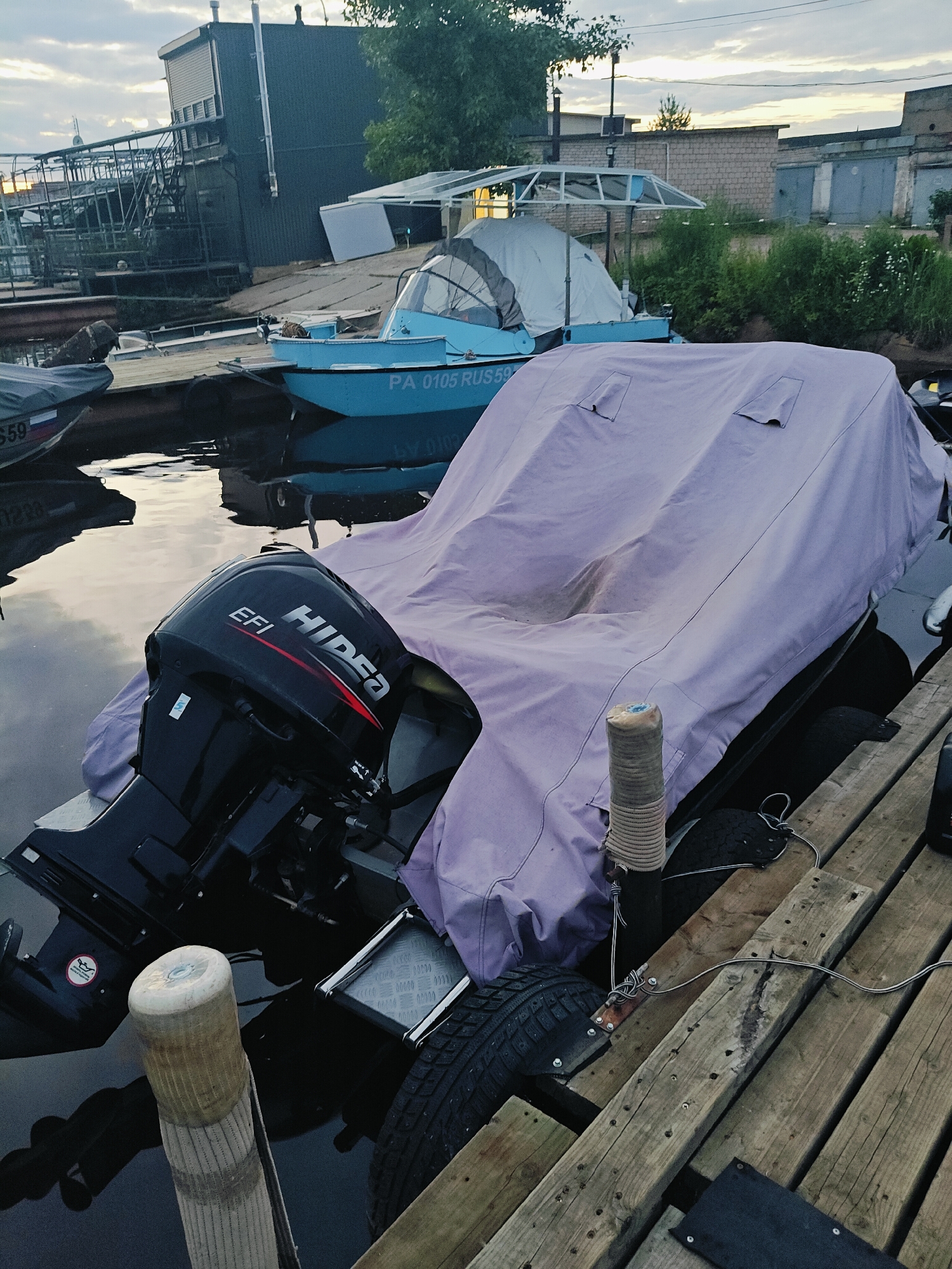 An overcast day at a marina in Chaikovsky, Russia, where several boats are docked. In the foreground, a large boat is covered with a light purple tarp. Its black outboard motor, with "HIDEN EFI" branding, is visible on the stern. A wooden pier runs diagonally across the right side of the frame, with a fender tied to a post on the left. In the background, a smaller blue boat with "PA 0105 RUSS" visible on its side is moored. Further back, industrial buildings and warehouses line the shore, with a cloudy sky overhead. The water is calm, reflecting the muted tones of the sky and surrounding structures.