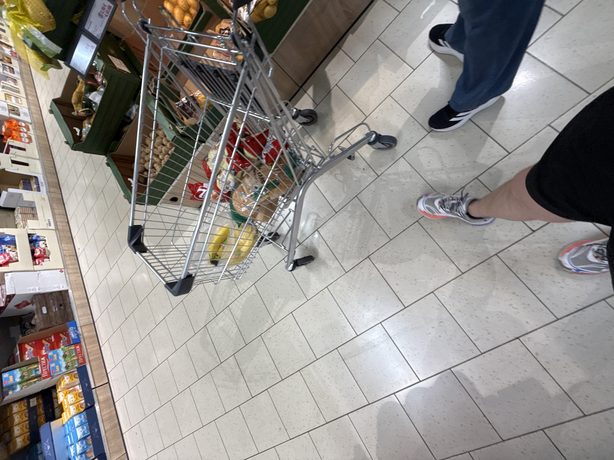 The image depicts a perspective from a person's point of view, looking down at their feet and a shopping cart in a grocery store. The floor is tiled with light-colored square tiles, some with scattered darker flecks. The shopping cart is made of metal and contains various grocery items, including bananas, bread loaves, and bags of chips or snacks. In the background, shelves stocked with packaged goods are visible, suggesting a retail environment. The lighting appears to be artificial, typical of an indoor store. The scene is consistent with a shopping trip in a modern supermarket, and could be located in Stara Zagora, Bulgaria, or any similar urban setting with such a retail establishment.