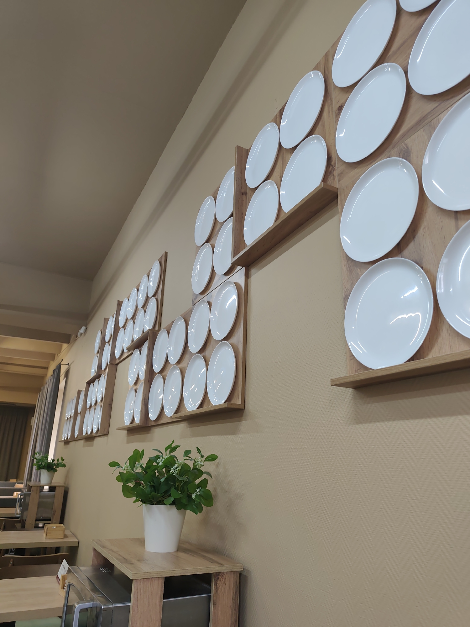 The image presents the interior of a dining establishment, likely a cafe or restaurant, located in Izhevsk, Russia. The central focus is a decorative wall adorned with numerous white ceramic plates of various oval and rectangular shapes. These plates are meticulously arranged in grid-like patterns on light wooden shelves and panels, creating a striking visual texture against the beige, textured wallpapered wall, which features a subtle herringbone pattern.
In the foreground and midground, several small green potted plants with delicate white flowers are placed in simple white pots on light wooden tables or low shelves. One prominent plant sits on a small wooden stand that also holds a metallic appliance, possibly a food warmer or dispenser.
The setting appears orderly and clean, with other wooden tables and chairs partially visible in the background, hinting at a seating area for patrons. The lighting is artificial and even, suggesting an indoor environment. No people are present, creating a quiet and static scene, possibly captured during off-hours or a lull in activity. There is no visible text in the image.