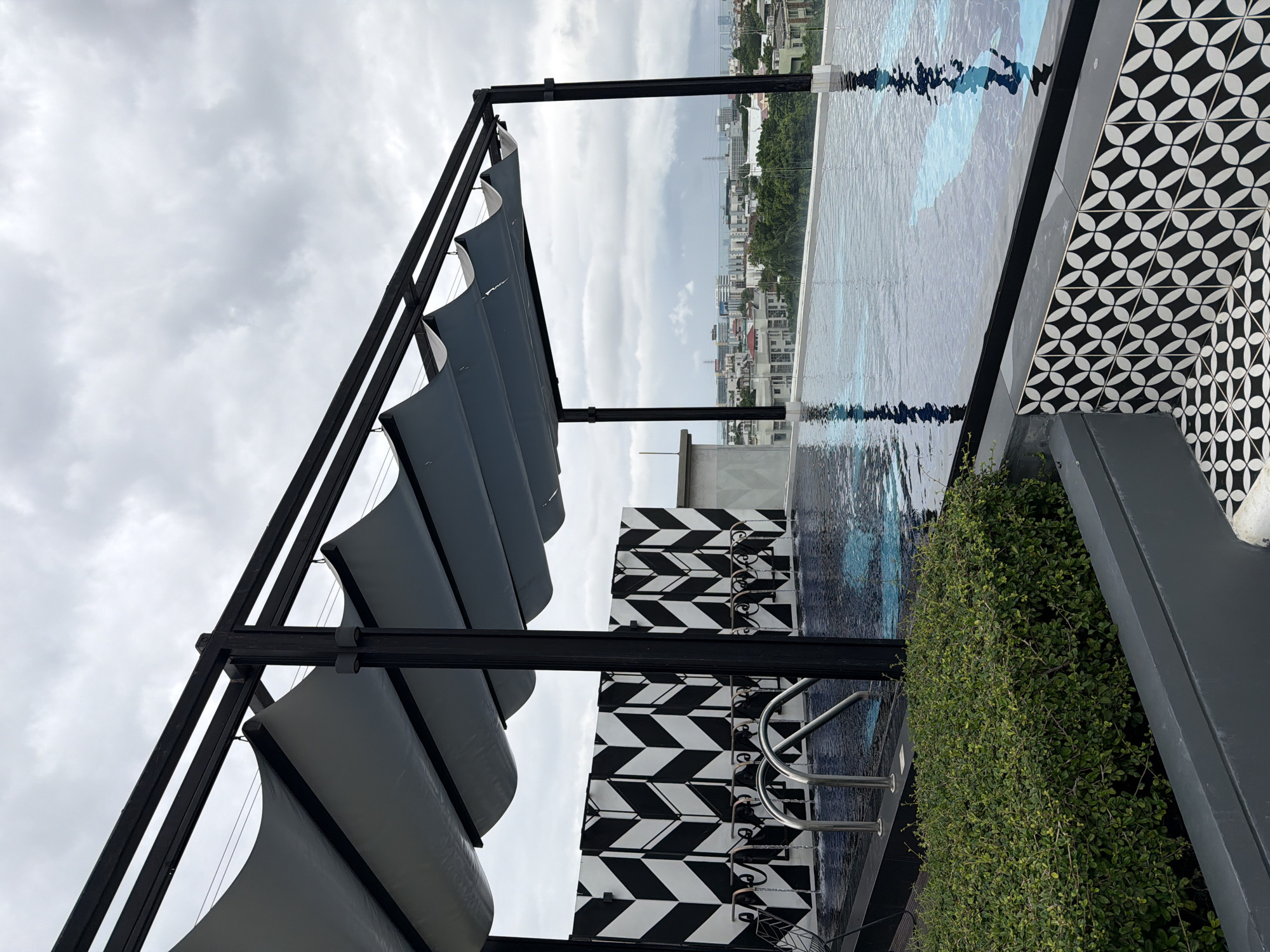 The image shows a rooftop infinity pool in Bangkok, Thailand. The pool is surrounded by a black and white patterned wall, with a green bush in the foreground. A black metal pergola with a fabric canopy covers part of the pool. In the background, the city skyline with buildings and trees is visible under a cloudy sky. There are no people in the image. The overall impression is one of a modern, stylish leisure space in an urban setting.