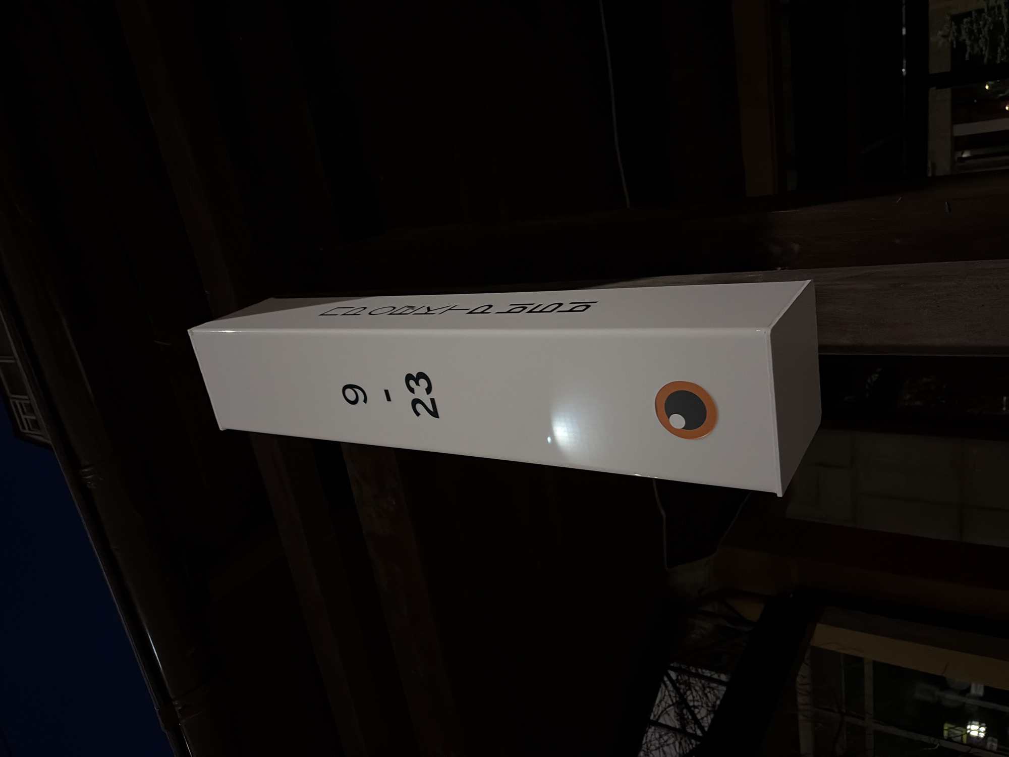 A white rectangular box with "COSMOS-D LTD" printed on top and "9-23" printed vertically on the side sits on a wooden beam. The box also features a stylized eye graphic on the visible end. The setting appears to be outdoors at night, possibly in an urban environment. The exact location in Perm, Russia, is not discernible from the given image. The focus is on the object and its immediate surroundings, revealing elements of the built environment, such as structural beams.