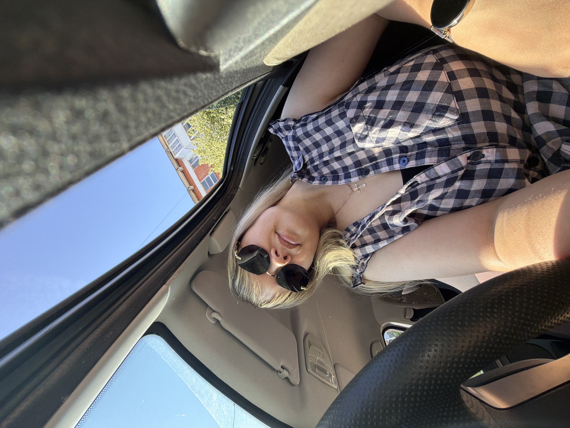 A low-angle view inside a car with a woman posing for the camera. The woman has blonde hair, wears sunglasses, and is dressed in a checkered shirt. Her facial expression is neutral. She wears a necklace with a cross pendant. Parts of the car's interior are visible, including the steering wheel, sun visor, windshield, and roof. The car appears to be parked outdoors, as the sky and a building can be seen through the windshield. The building visible outside the car has a light-colored facade and red brick accents. The location is likely in Russia, in the city of Chaikovsky.