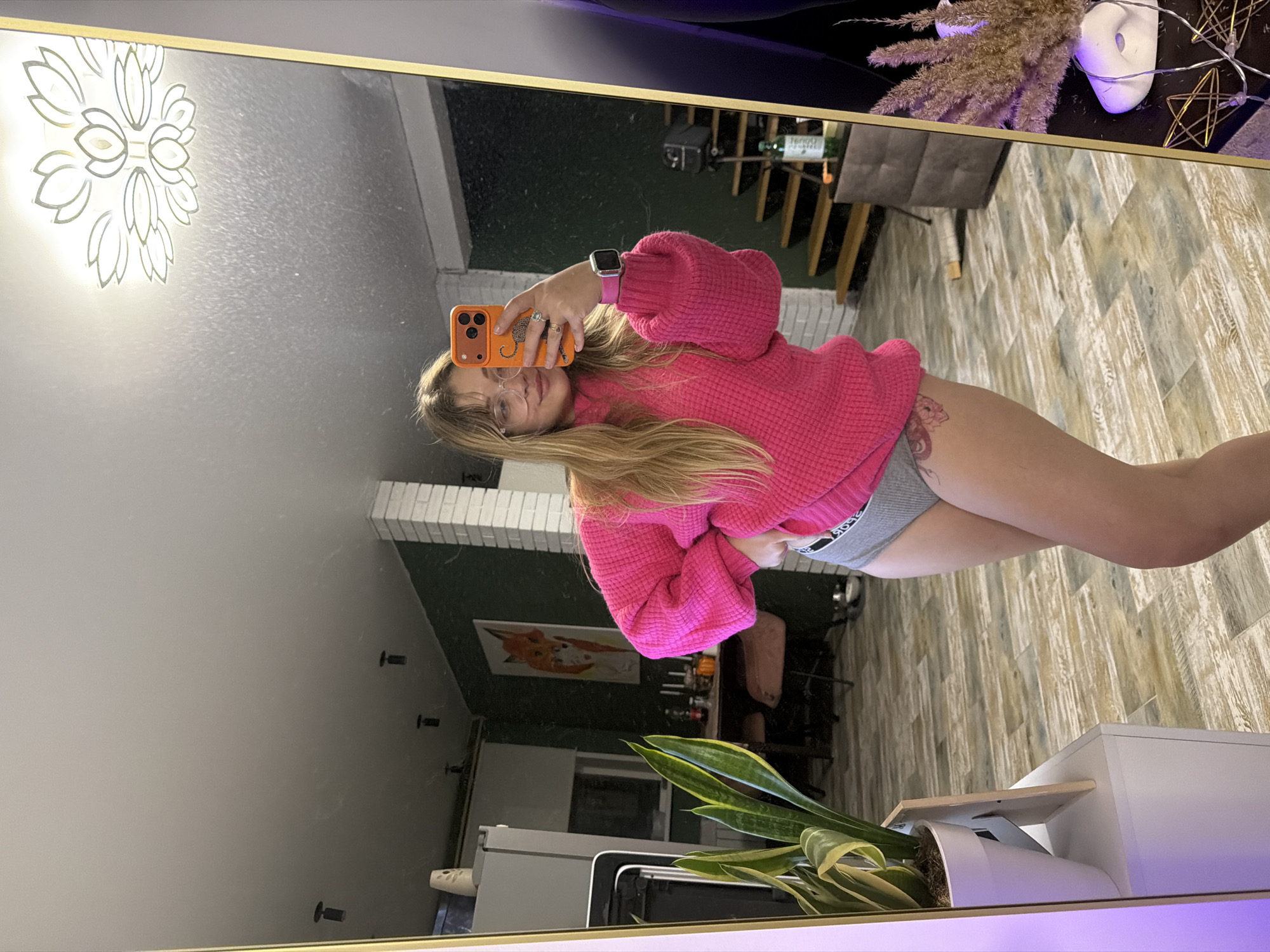 A woman takes a selfie in a mirror. The mirror is reflecting part of a room in a building located in Большая Шильна, Russia. The room contains an indoor plant, a table, a chair, and a painting of a fox. The woman is wearing a pink sweater.