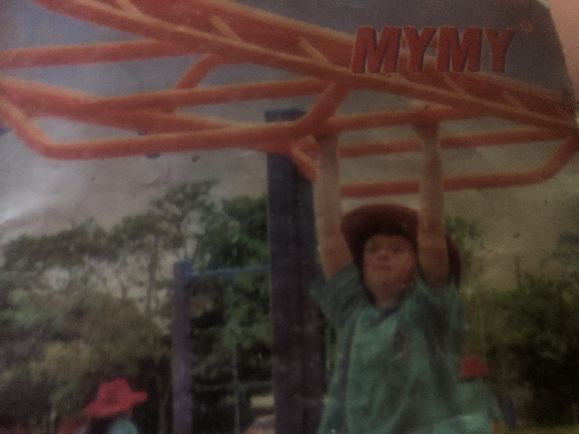 The image shows a person lying down, wearing a green garment and a cowboy hat. They are positioned on a dark surface, possibly a bench, with a railing to their right. The background is blurred and appears to be an outdoor setting with greenery and some orange structures. There is a red object visible to the left of the person. Text reading "MYMY" is present vertically on the orange structure. The image is of poor quality, with significant blur and noise. The location in Wukari, Nigeria cannot be determined from the provided image.