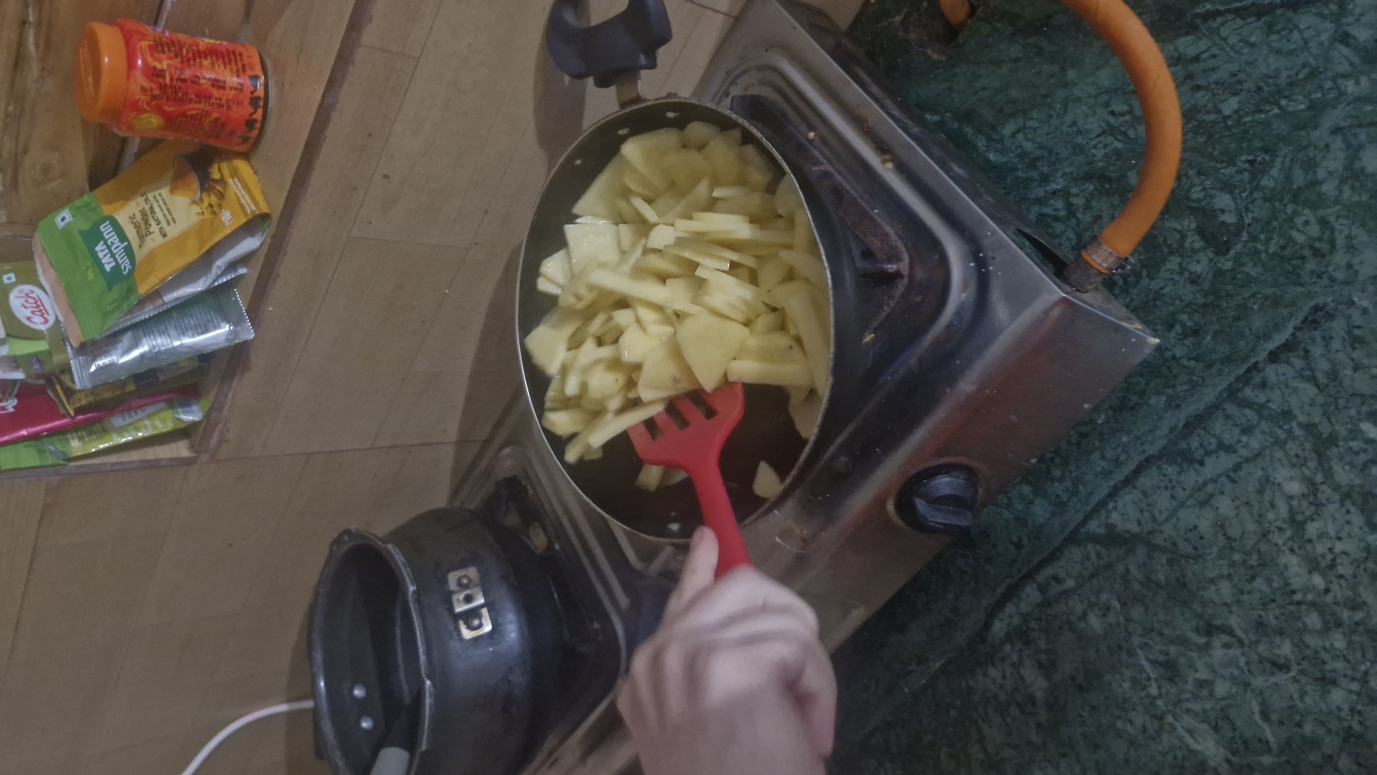 In an indoor kitchen setting, a close-up shot captures a hand holding a red spatula, stirring chopped potatoes in a frying pan on a gas stove. The potatoes are uniformly cut and appear to be raw. To the left of the frying pan, a black pressure cooker is visible on the stove. In the background, shelves are lined with various packaged spices and condiments, including a pack of "TATA Sampann Turmeric Powder" and a red spice jar. The overall environment suggests a home kitchen with a focus on food preparation.