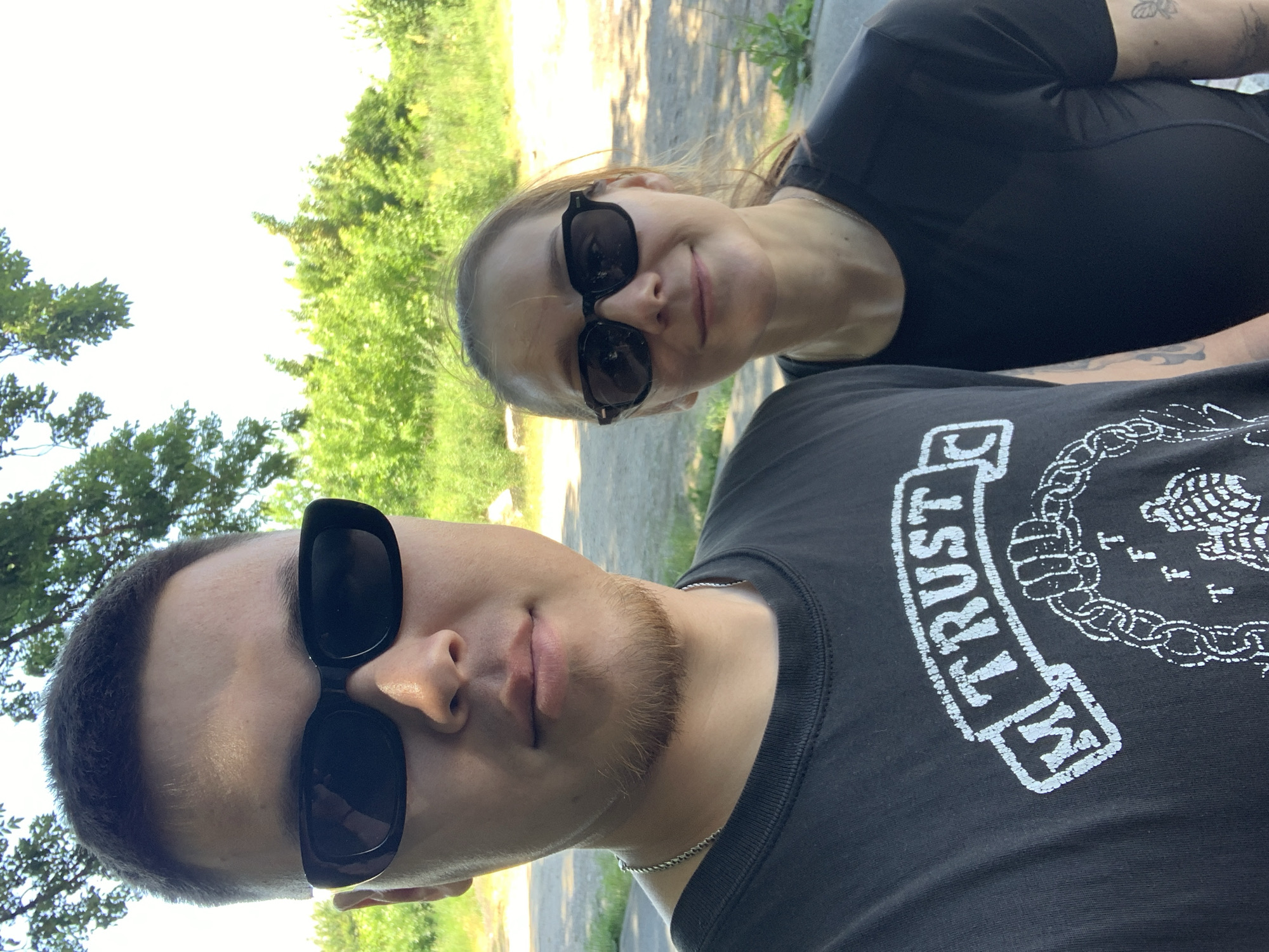 This is a close-up, vertical, outdoor selfie of two young people, a man and a woman, who appear to be in their early twenties. They are both wearing dark-colored t-shirts. The man, in the foreground, is wearing sunglasses and a silver chain necklace. He has short, dark hair and a light brown goatee. The woman, behind him, is also wearing dark sunglasses and has blonde hair. They are smiling slightly. The setting appears to be a park or wooded area, with lush green trees and dappled sunlight visible in the background. The ground looks like a dirt path. The time of day is likely daytime due to the bright lighting and shadows. The image has a casual, relaxed feel, suggesting a friendly outing. There is visible text on the man's t-shirt, partially obscured, which appears to be a distressed white graphic. The words "M TRUST" are clearly legible, within a box-like design. The location is Stavropol, Russia.