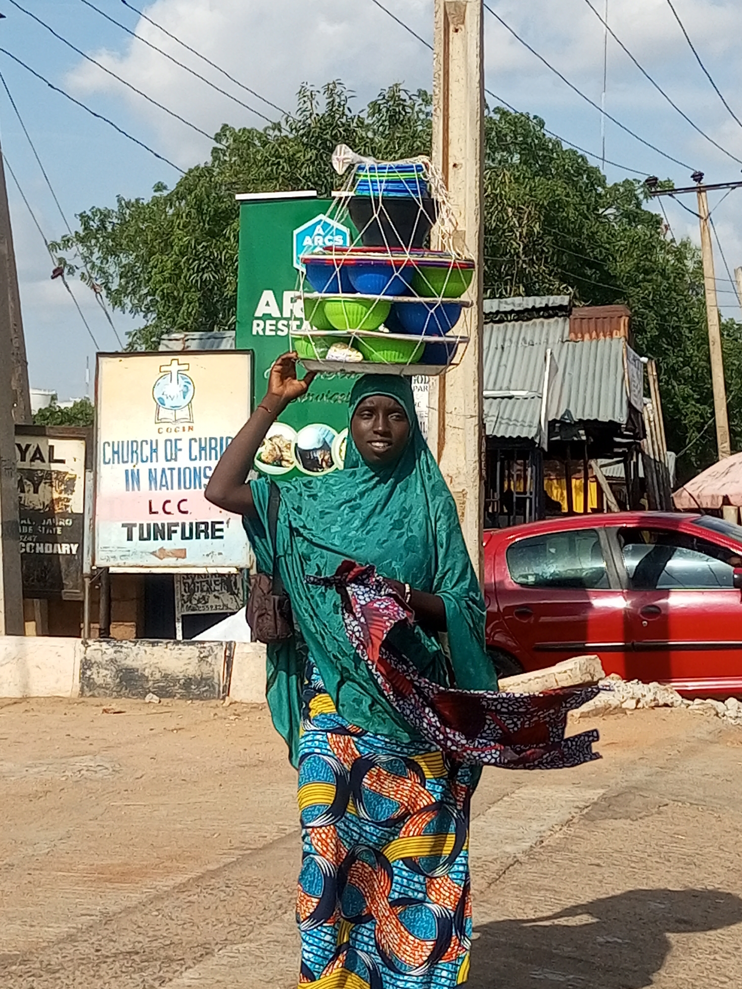 A young woman, appearing to be in her late teens or early twenties, is walking outdoors in Gombe, Nigeria. She is wearing a green hijab and a brightly colored, patterned wrap skirt. On her head, she carries a tiered stack of bowls, secured with netting and rope. In her right arm, she is holding a folded piece of fabric with a red, white, and blue pattern.
The scene appears to be on a dusty, unpaved street with some concrete elements. Behind the woman, there are several signs. One prominent sign, with a green background, features the logo of "ARCS" and text that reads "RESEARCH". Another sign, cream-colored, displays a cross and an open book, with text identifying it as "CHURCH OF CHRIST IN NATIONS LCC TUNFURE". Further signs with text like "YAL" and "CCOARY" are also visible.
To the right of the woman, a red car is parked. The background includes trees and a partially visible structure with a corrugated metal roof. Overhead, power lines crisscross the sky, which is partly cloudy. The overall impression is of a typical street scene in a Nigerian city, likely during the daytime.