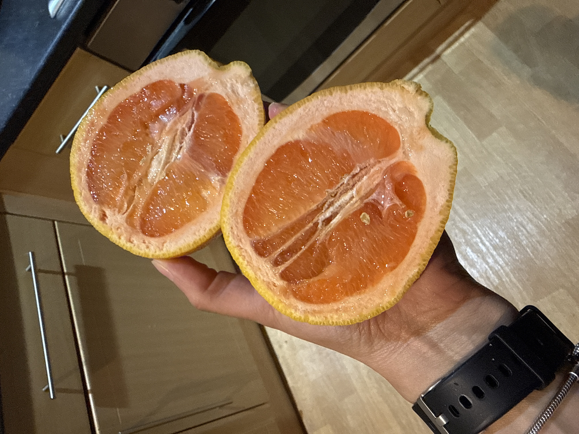 A hand holds a halved grapefruit indoors. The grapefruit has a yellow rind and pinkish-orange flesh. A black watch is on the wrist. The background consists of wooden cabinets. There's no evidence of this image being taken in Great Yarmouth, United Kingdom.