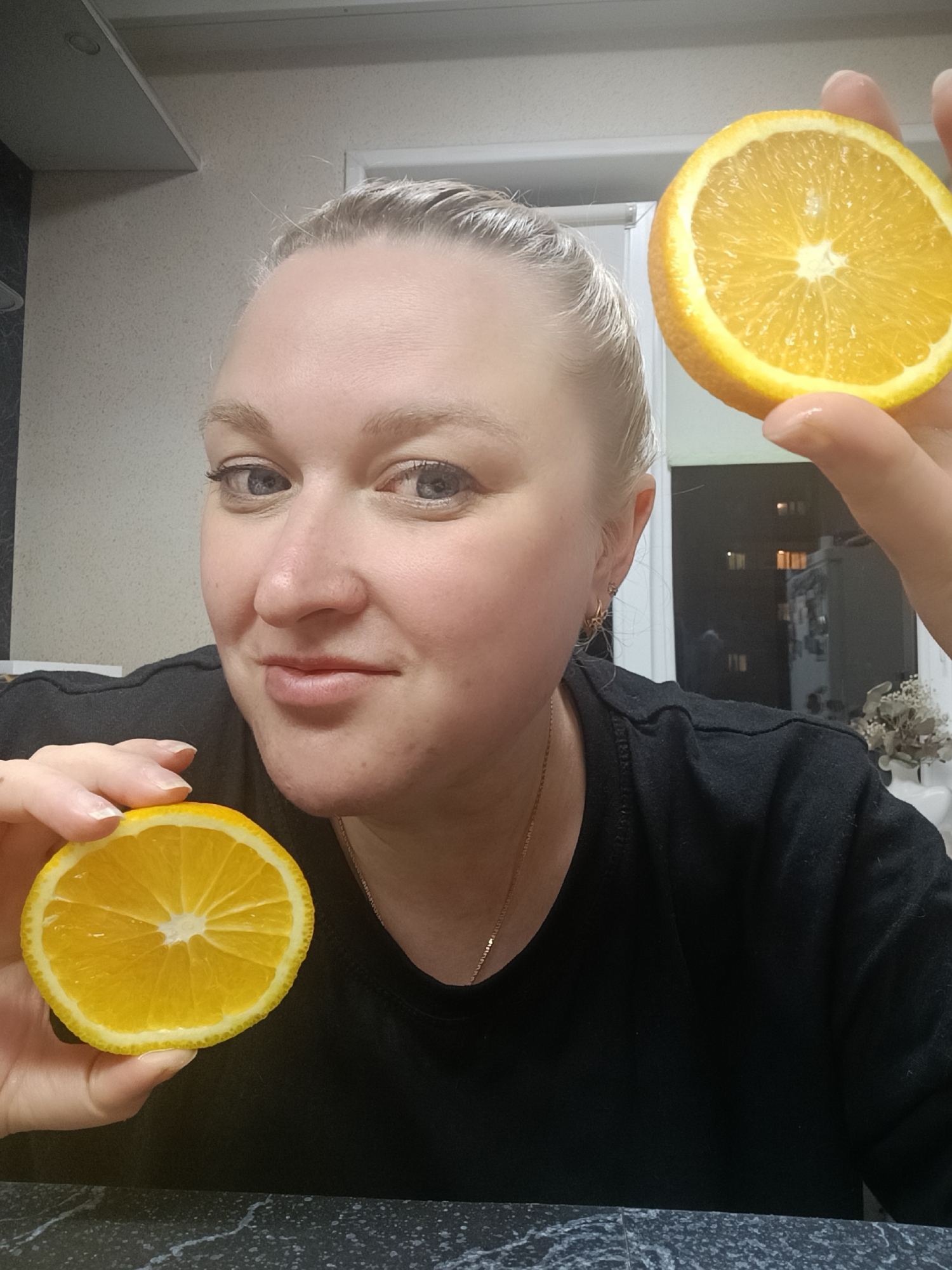 A fair-skinned, blonde woman is holding two orange slices in front of her indoors. She is wearing a black shirt and a gold necklace. The background shows a kitchen setting with dark countertops and light-colored cabinets. Outside a window, there's a view of an urban environment with visible buildings and illuminated windows, suggesting it may be evening. The location is consistent with an urban setting, but pinpointing the exact spot in Chaikovsky, Russia without additional contextual clues is not possible.