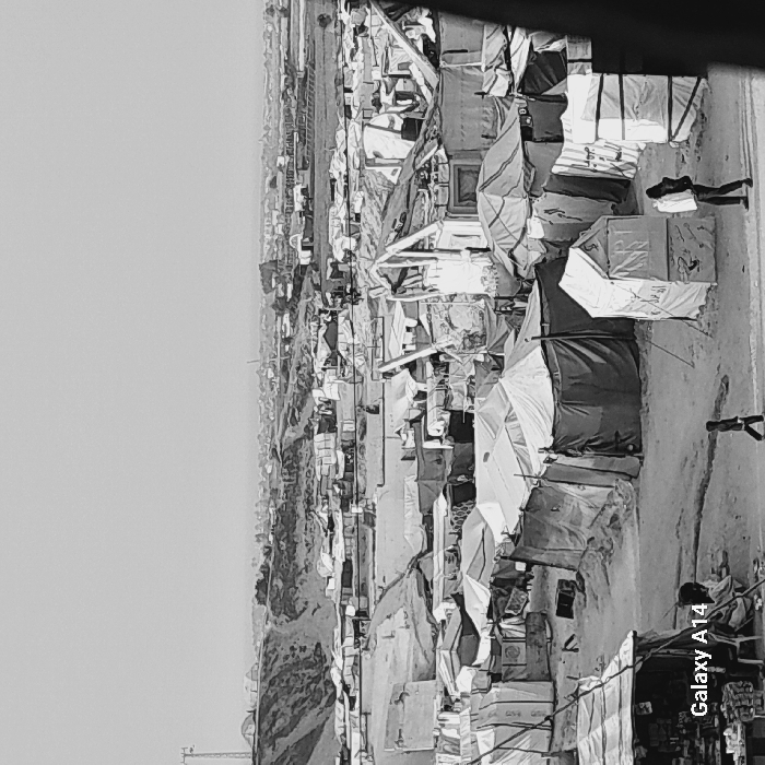 The image displays a densely populated makeshift encampment in Khan Yunis, Palestinian Territory. Hundreds of temporary shelters, primarily constructed from tarpaulins, fabric, and other salvaged materials, are tightly clustered across an unpaved, sandy, and uneven landscape. In the left background, a large dirt hill is visible, topped with a communication tower, with a broader urban or agricultural panorama extending into the hazy distance. In the foreground, a rudimentary stall displaying goods is visible at the bottom left, near a seated individual. A small child walks leftward through a cleared path in the lower-center. On the lower right, an adult figure in dark clothing walks rightward, carrying a large, light-colored rectangular object. No direct interactions between the visible individuals are apparent.