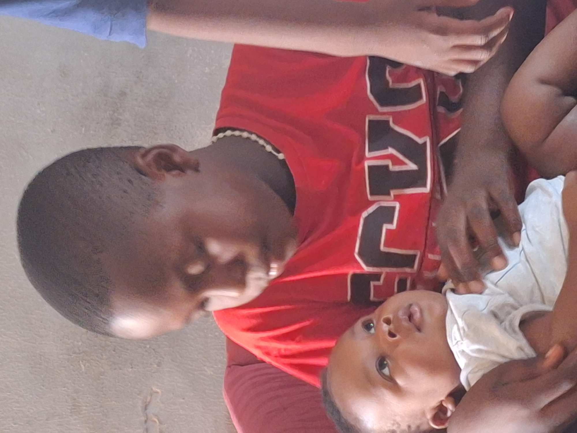 A tilted, close-up image shows an adult holding a baby, likely indoors in Sabon Tasha, Nigeria. The adult has short dark hair and wears a red short-sleeved shirt with large, partially visible block letters on the front and a light-colored beaded necklace around their neck. The adult's head is tilted downwards, and they are looking towards the baby with a gentle expression. The baby, dressed in a light-colored top, is looking upwards. An adult's hand is visible resting on the baby's body, suggesting a moment of care and connection. The background is a plain, light-colored wall.