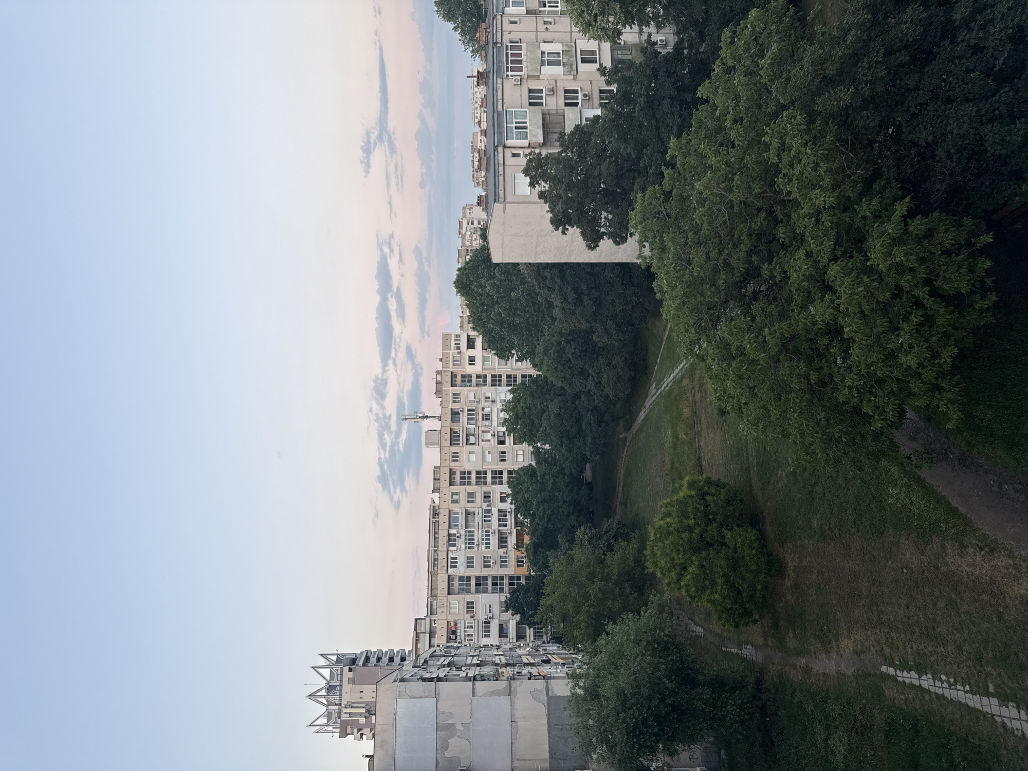 The image presents an urban vista in Plovdiv, Bulgaria, captured during a period of low-angle sun, indicated by a sky transitioning from light blue at the zenith to pale pink and orange near the horizon, with scattered light grey stratocumulus clouds. The perspective is elevated, overlooking a residential area featuring a central green space.
In the left half of the frame, a multi-story building, identified as the Grand Hotel Plovdiv (formerly Trimontium Hotel), rises prominently with a distinctive, multi-pronged crown-like structure on its roof, which hosts several communication antennas. Immediately in front of it is a weathered, multi-story concrete residential building, gray with exposed aggregate and peeling plaster, featuring numerous windows, enclosed balconies, and attached exterior air conditioning units. A series of similar, densely packed apartment blocks extends into the background.
Horizontally spanning the middle ground is a substantial light-colored apartment block, also characterized by a grid of windows, multiple balconies, and visible air conditioning units. Another grey apartment building is partially visible on the far right. The lower half of the image is occupied by a park-like green area, densely vegetated with mature deciduous trees, exhibiting dark green foliage. Unpaved dirt paths meander through the grass; one distinct path in the lower left is delineated by light-colored paving stones and runs alongside a low wall bearing faint graffiti. No individuals or vehicles are present within the observed scene.