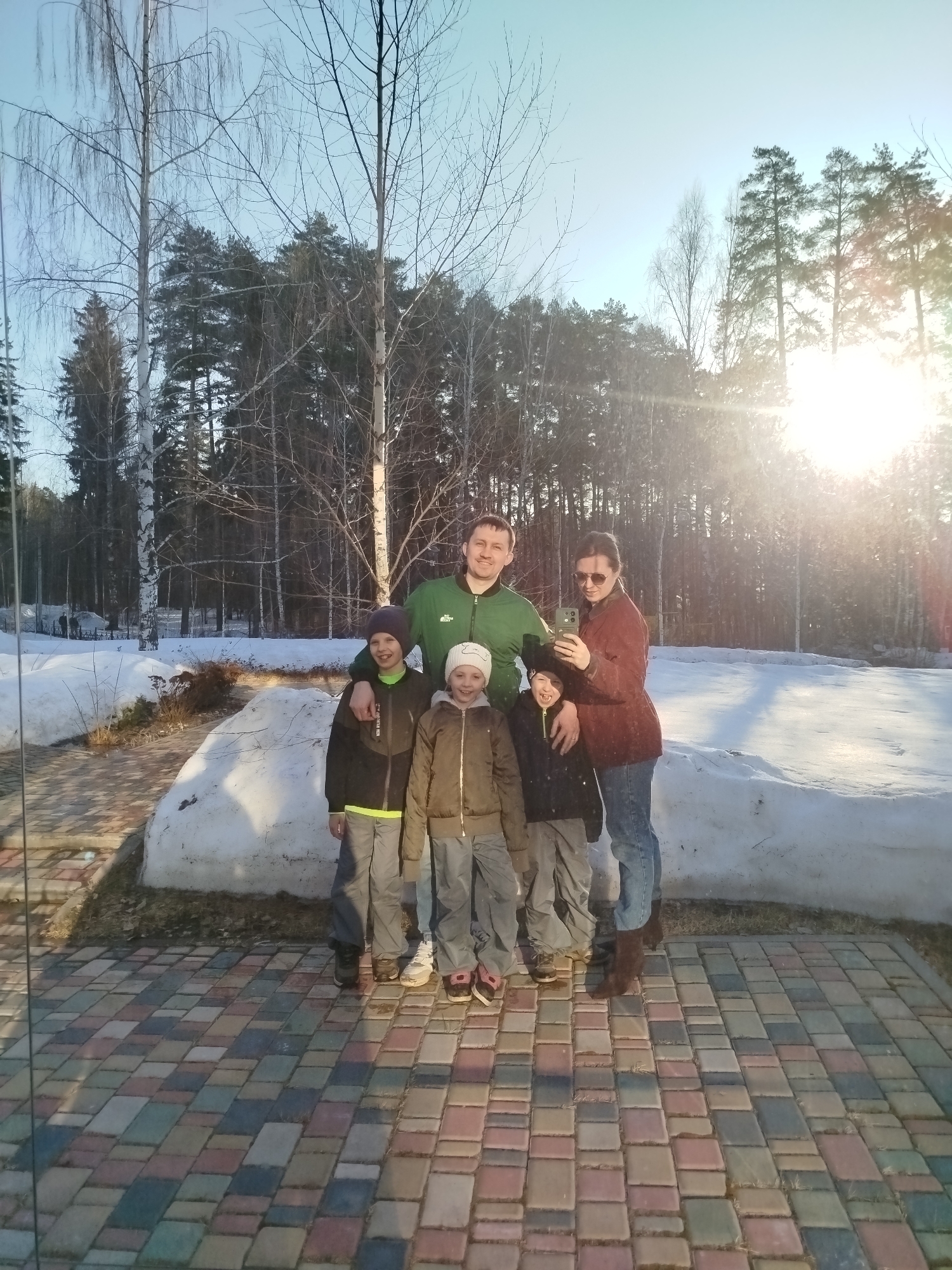 A group of five people stands in front of a large snow pile on a bricked area, with a backdrop of trees and a bright sun.
The people are comprised of two adults and three children. The man is wearing a green jacket and has his arms around two of the children. The woman is wearing a red jacket and sunglasses. The children are dressed in winter coats and hats.
The setting appears to be outdoors, potentially in a park or a yard in Izhevsk, Russia. Remnants of snow are on the ground. A forest of trees fills the background. The sky is blue.