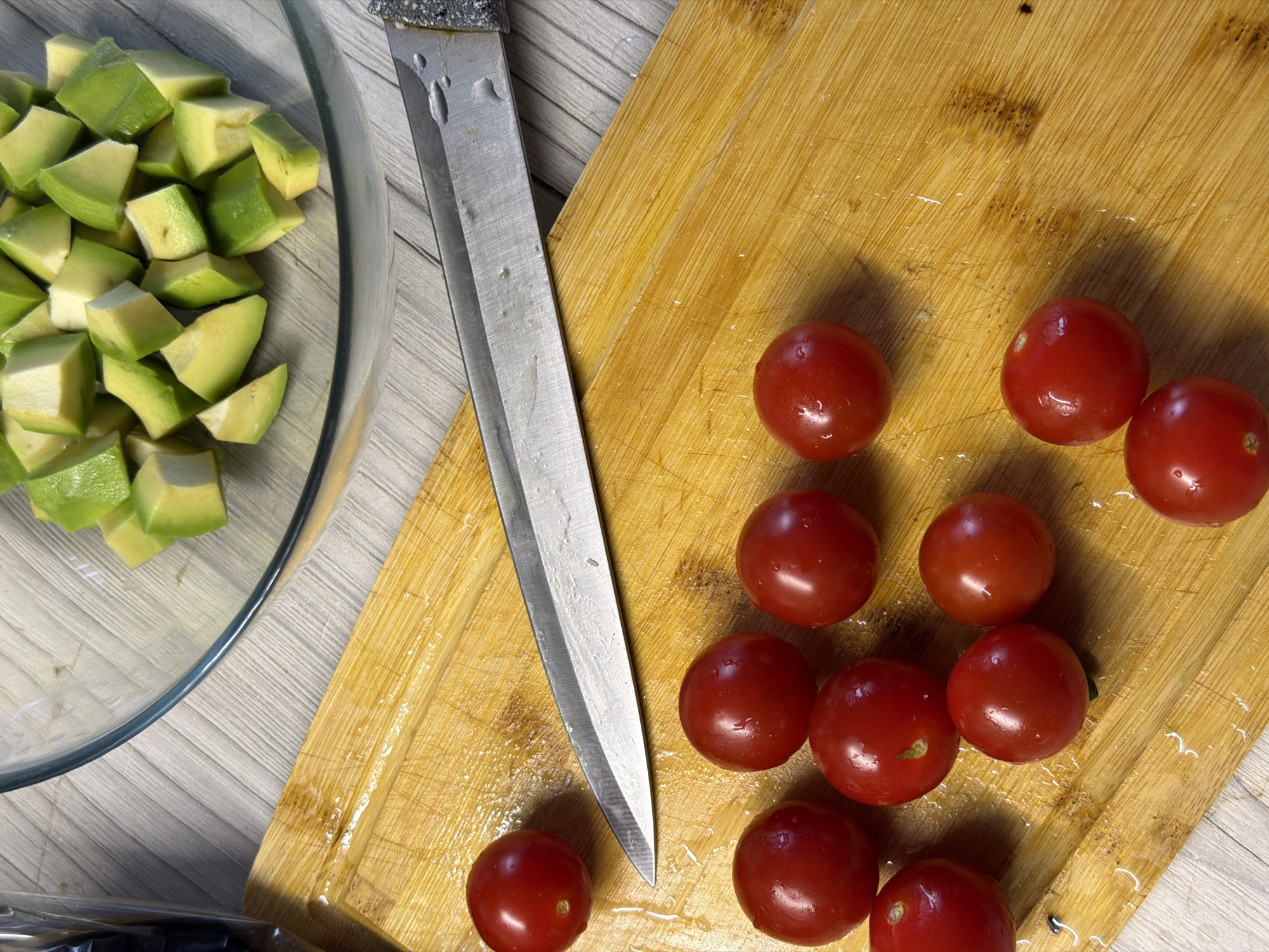 The image shows a close-up, overhead view of a wooden cutting board with several ripe cherry tomatoes scattered across it. A metal knife with a dark handle lies diagonally across the cutting board, with its blade pointing towards the bottom left corner of the image. To the upper right of the cutting board is a clear glass bowl filled with diced avocado. The background consists of a light-colored, textured surface, possibly a countertop or table. There is no indication of the city of Chaikovsky, Russia, or any people in the image.