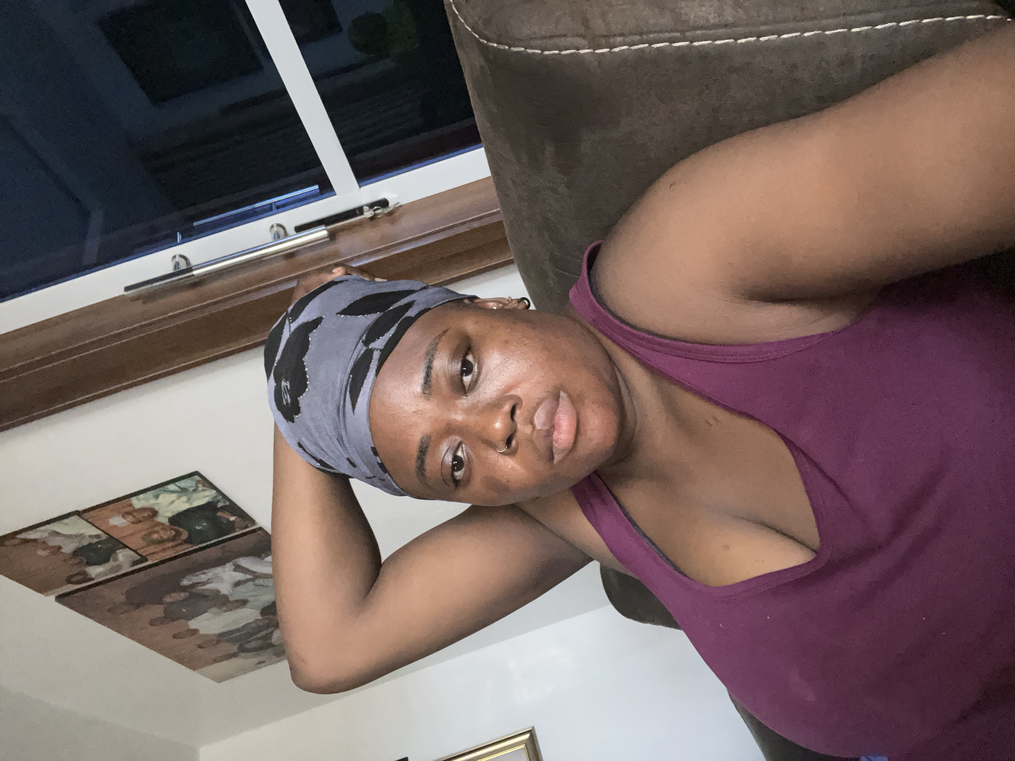 A woman with brown skin, wearing a purple tank top and a gray and black headscarf, is lying down, facing the camera. Her left arm is bent behind her head. She has a nose piercing. In the background, there is a window with a wooden frame, a brown leather couch, photos on the ceiling, and a gold-framed object. The location and city are not discernible from the image.
