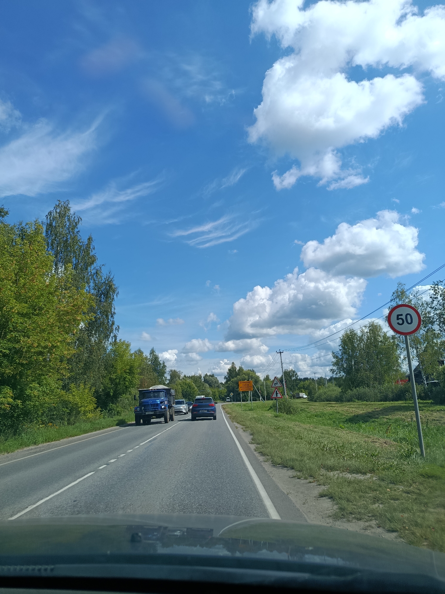 The image shows a view from a vehicle driving on a road near Vereya, Russia. A blue truck is in the foreground, followed by other cars. The road is lined with trees and a grassy verge. A speed limit sign of 50 is visible on the right side of the road. The sky is blue with white clouds. There are also road signs visible, including one that appears to warn of children crossing. The scene suggests a sunny day in a rural or suburban area.