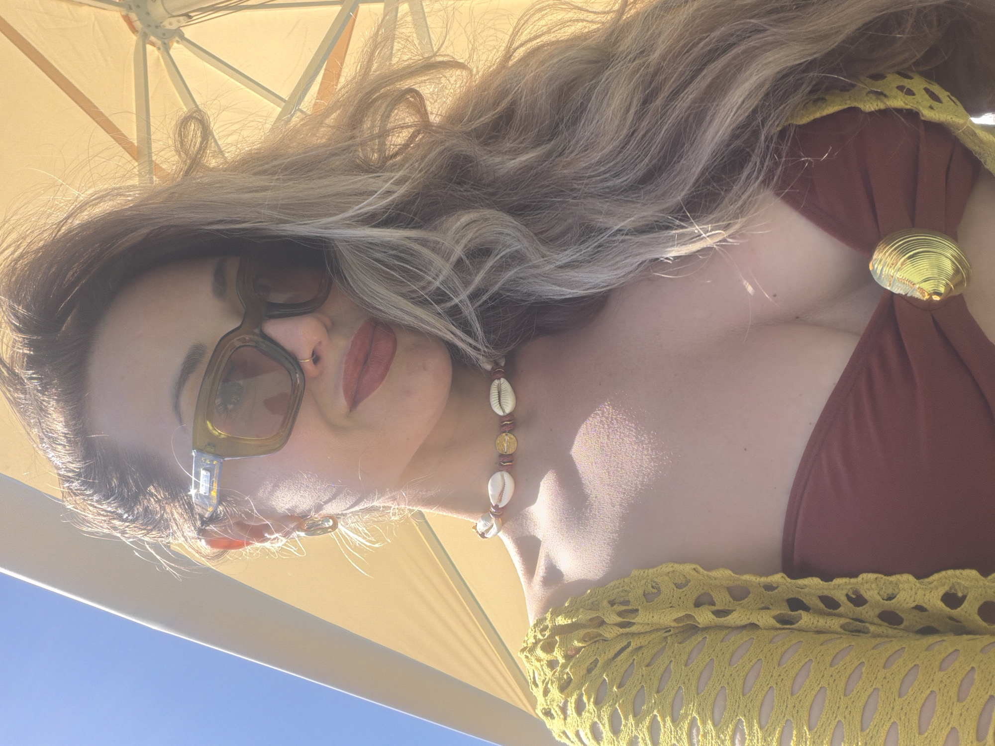 A close-up portrait shows a woman relaxing outdoors, viewed from a slightly low angle. The woman has long, wavy light brown hair. Her face is partially obscured by rectangular-framed sunglasses with dark lenses and light green/brownish frames. She wears distinct red lipstick and a small gold ring piercing in her nose.
She is dressed in a dark red or maroon bikini top, featuring a prominent gold shell-shaped ornament on the strap near her cleavage. Over the bikini, she wears a loosely crocheted or fishnet-patterned yellow cover-up. Around her neck, she has a necklace made of white cowrie shells interspersed with brown beads and small gold-colored disc beads.
The background consists primarily of the light yellow or beige underside of an umbrella or awning, providing shade. Below this structure, a clear blue sky is visible. The bright sunlight filtering through the umbrella and illuminating her hair suggests it is daytime. The overall scene conveys a sense of leisure and warmth. There is no visible text in the image. The context provided for this image is Krasnoyarsk, Russia.