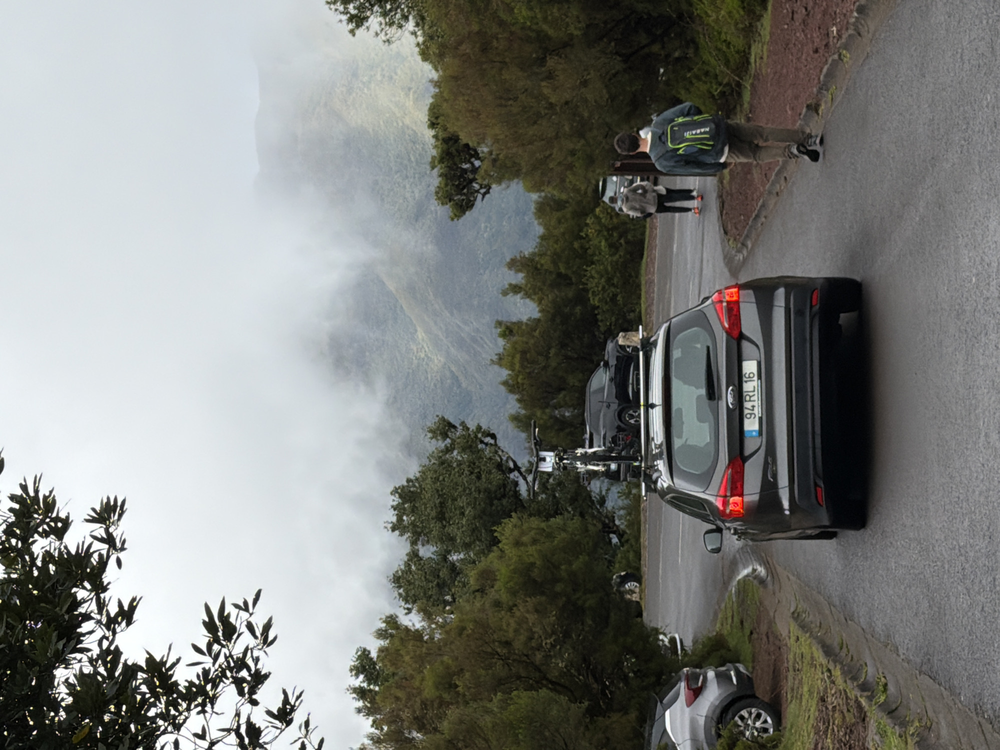 The image shows a street scene with a mountainous backdrop in Porto Moniz, Portugal. Fog obscures the view of the peaks. A dark gray car with license plate 94 RL 16 is parked on the right side of the road. Behind it, a bicycle is visible. Two people are walking along the road, one of whom is wearing a backpack with the text "NABAIJI" on it. Another car, lighter in color, is parked behind the gray one. The road is wet, and there is greenery including trees and shrubs lining the roadside. The sky is overcast.