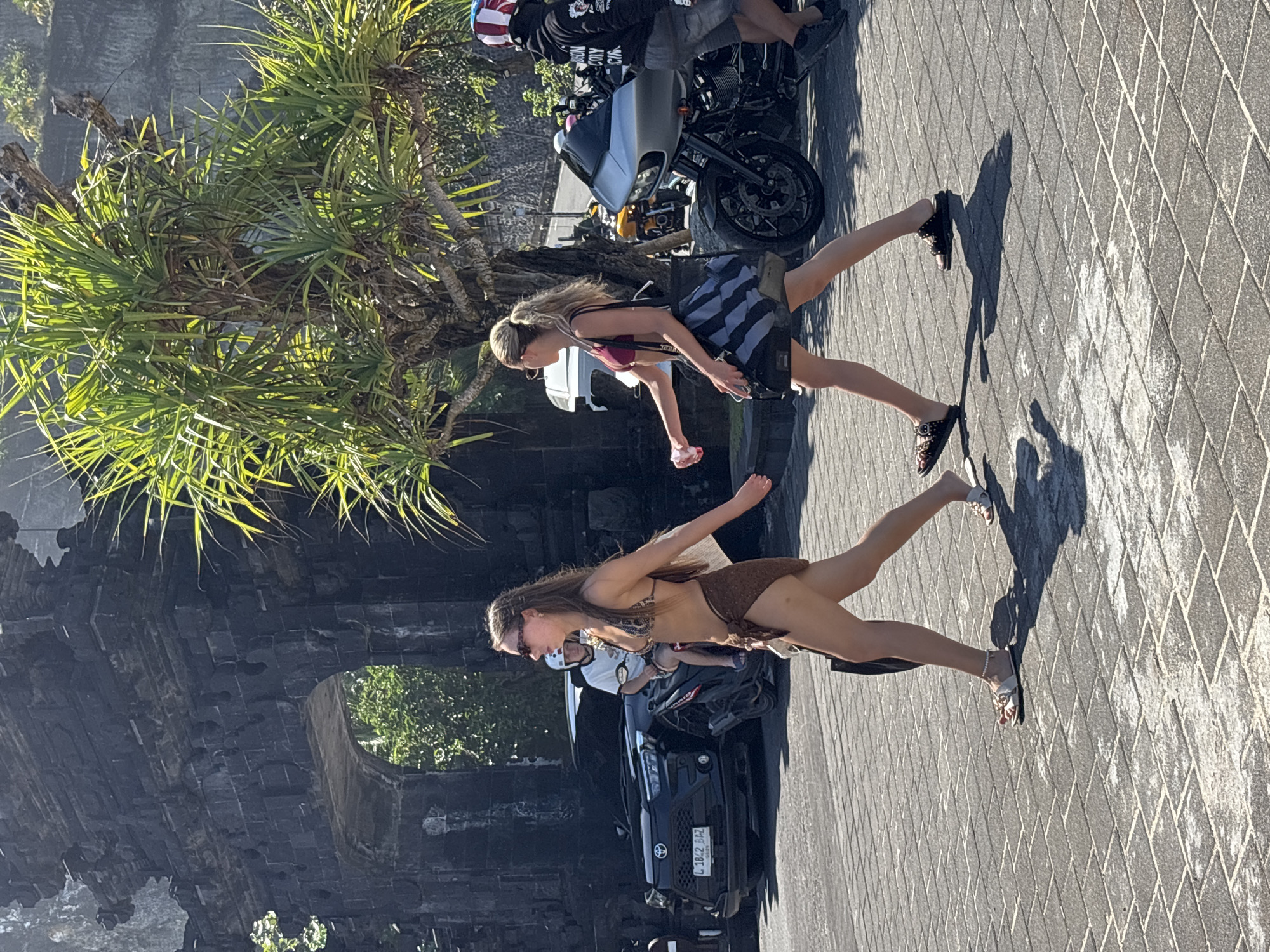 In Kuta Selatan, Indonesia, two women walk on a paved walkway in front of a large, dark stone archway. The woman on the left wears a brown and black patterned bikini, a brown sarong, and white sandals, and carries a dark bag. The woman on the right wears a maroon bikini top and a black and grey striped skirt, and carries a dark bag. Behind them, a black car with a license plate ending in "BAZ" is parked. A motorcycle with a silver front fairing is parked to the right of the women. A person wearing a helmet with red, white, and blue stripes stands next to the motorcycle. Green foliage from a tree is visible in the upper right corner of the image. The ground is paved with grey rectangular stones.
