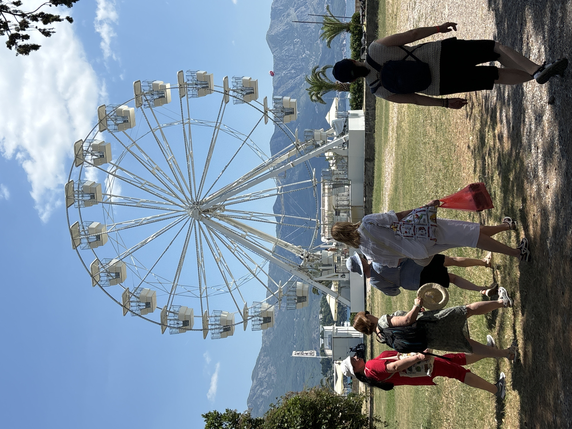 A large white Ferris wheel stands prominently against a clear blue sky with sparse clouds, overlooking a coastal landscape with distant mountains. In the foreground, a group of five women walks away from the camera on a gravel path with patchy green grass. They are dressed casually for warm weather, with some carrying bags and one holding a hat. The Ferris wheel appears to be in operation, with several of its gondolas visible. The overall scene suggests a sunny day at a seaside amusement park or promenade in Budva, Montenegro, given the distinctive mountainous coastline characteristic of the region.