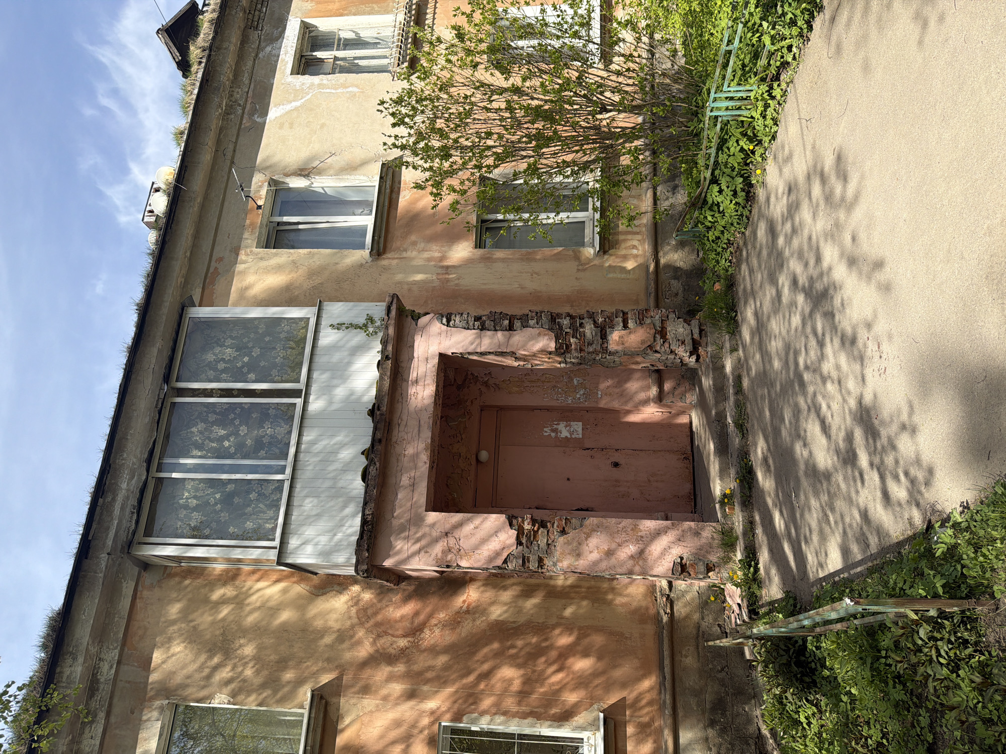 An exterior shot shows a two-story dilapidated building. The façade features a damaged, pink-painted entryway with a wooden door. Parts of the surrounding brickwork and plaster are crumbling. Above the entryway is a balcony or window with floral curtains. The building's paint is peeling. Vegetation, including a small tree, grows adjacent to the building and the adjacent sidewalk. Grass grows on the roof. A rusted metal fence borders the sidewalk. The location is Tokkari, Russia.