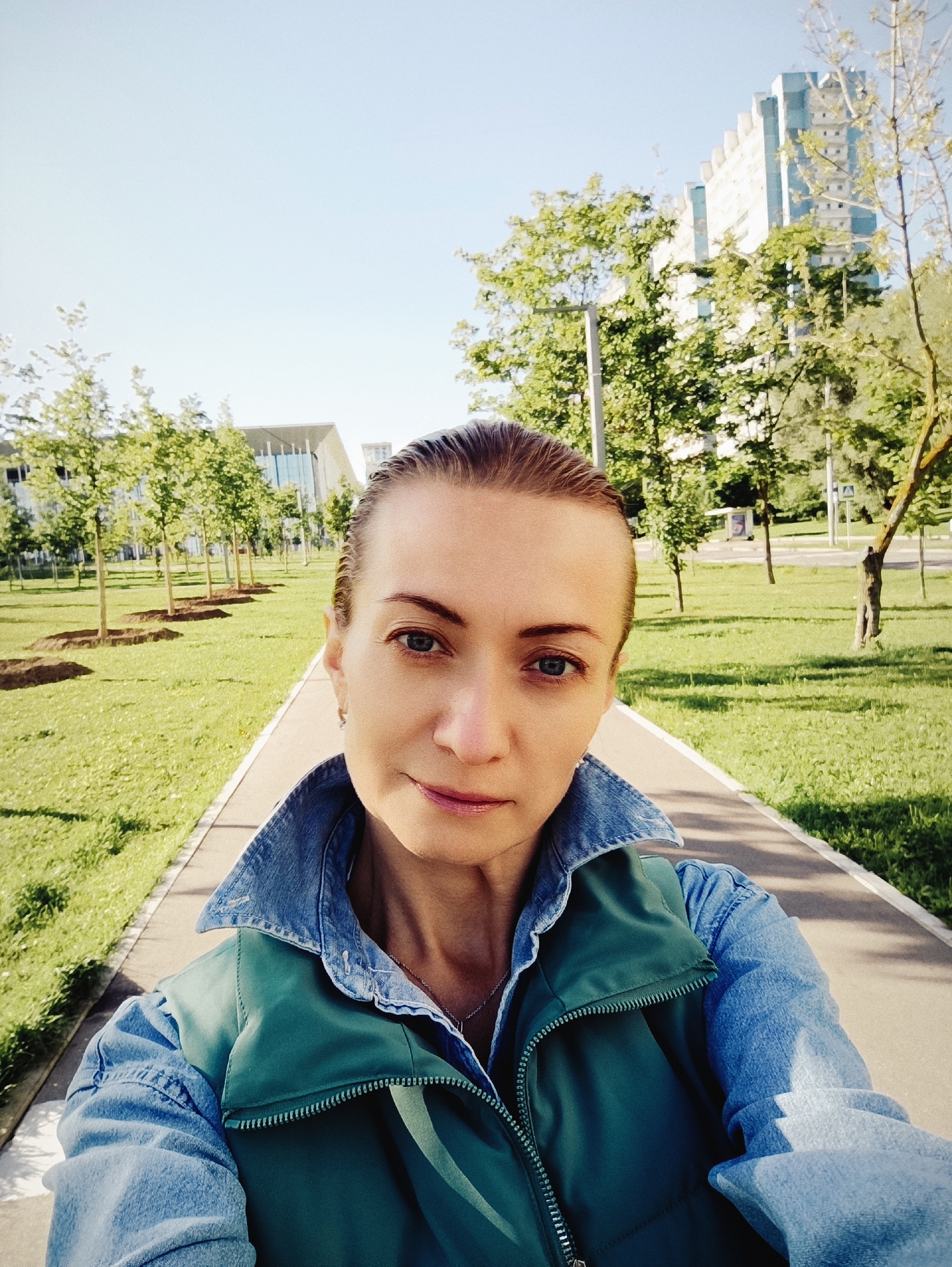 In a selfie taken outdoors, a woman with her hair pulled back smiles subtly as she looks at the camera. She is wearing a denim shirt with a green vest over it. Behind her, a paved walkway runs through a park-like setting with manicured trees and grass. In the background, several modern buildings are visible, including a tall, light-colored building with blue accents. The scene suggests an urban environment, likely a park or public space in Moscow, Russia, during daylight.
