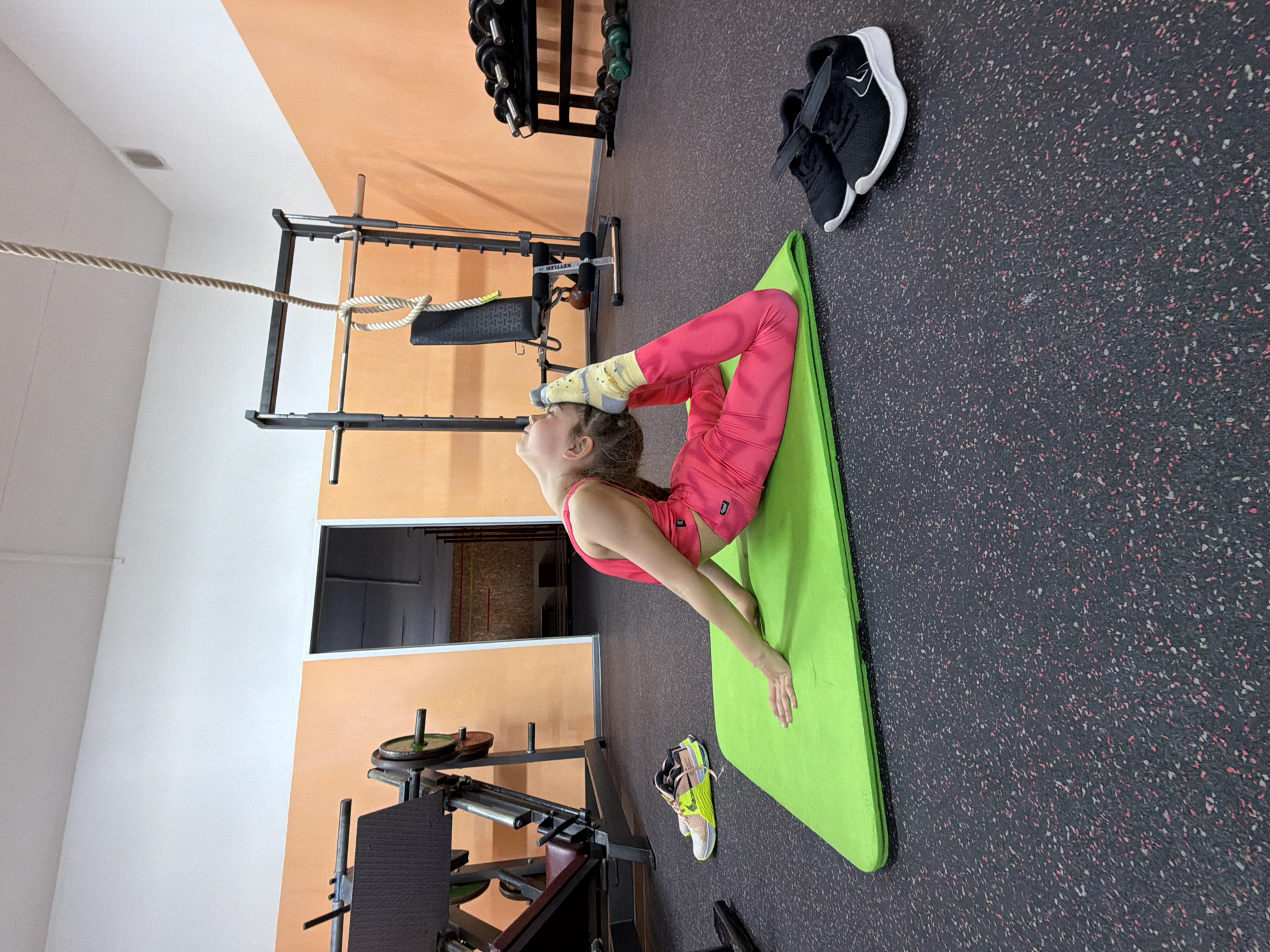 A girl is practicing a yoga pose, bending backwards, on a green yoga mat inside a gym. She is wearing pink athletic leggings and a matching tank top, with patterned socks covering her feet, which are arched over her head.
The gym has a black and red speckled floor. Several pieces of weightlifting equipment are visible, including a squat rack with weights, and a rope hanging from the ceiling. Two pairs of athletic shoes are near the mat.
The location is implied to be in the city of Chaikovsky, Russia.