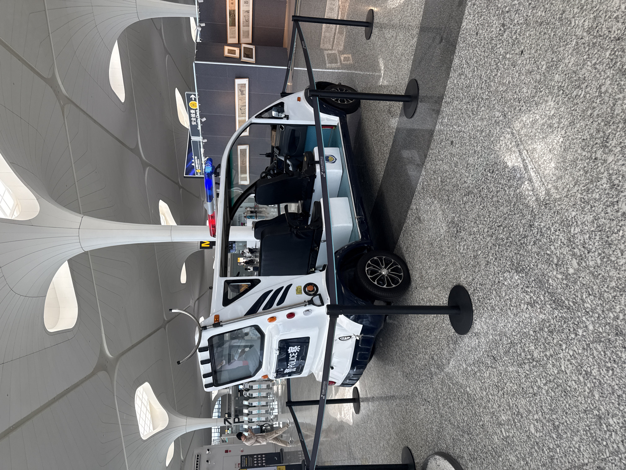 In the spacious, modern interior of Xiaoshan District's airport terminal, a white and blue electric police cart is displayed. The cart, equipped with flashing red and blue lights on its roof, features "POLICE" in prominent lettering on its rear. It is cordoned off with black stanchions and ropes, suggesting it is on display or awaiting service. In the background, airport check-in counters are visible, along with a person walking and signs pointing to "Security Check." The ceiling architecture is characterized by undulating white panels and structural columns, indicative of contemporary airport design. The floor is tiled with a speckled granite pattern.