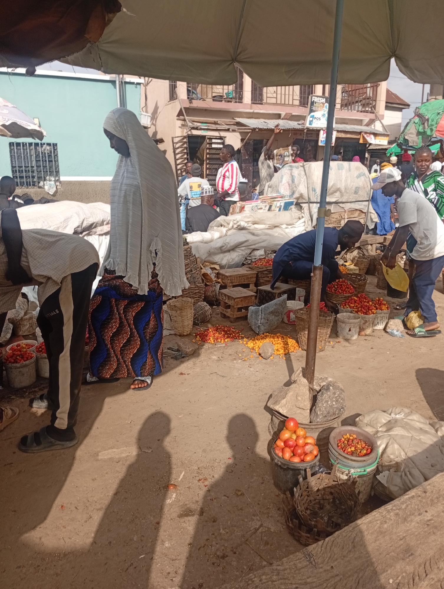 The image depicts an outdoor marketplace scene, likely in Rafin Par, Nigeria. The ground is dirt, with a few scattered pebbles and debris. In the foreground, a person wearing black and white striped pants and black sandals is bending over a bucket of red tomatoes. To their right, a woman in a light grey headscarf and a patterned blue and brown skirt stands near a stall.
Further into the scene, a man in a dark blue outfit is seen crouching and arranging baskets filled with red and yellow peppers. Another man, wearing a white shirt and blue pants, stands nearby holding a yellow plastic bag. In the background, several stalls are visible, some covered with tarps, with various goods, including what appear to be sacks of produce and other items. A few people are scattered around, some in the background appearing to be vendors or shoppers. The overall atmosphere suggests a bustling local market.