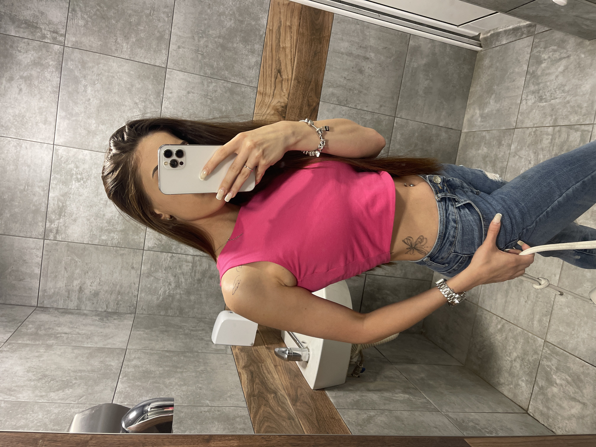 In the lavatory of an unknown establishment in an unknown city, a young woman with long brown hair is taking a selfie in a mirror. She is wearing a bright pink cropped tank top, distressed blue jeans, and silver jewelry. A small butterfly tattoo is visible on her left hip, and a subtle wing tattoo adorns her left shoulder. She holds a white smartphone in her right hand, capturing her reflection. The bathroom features grey tiled walls and floor, a white sink with a silver faucet, and a silver hand dryer mounted on the wall.