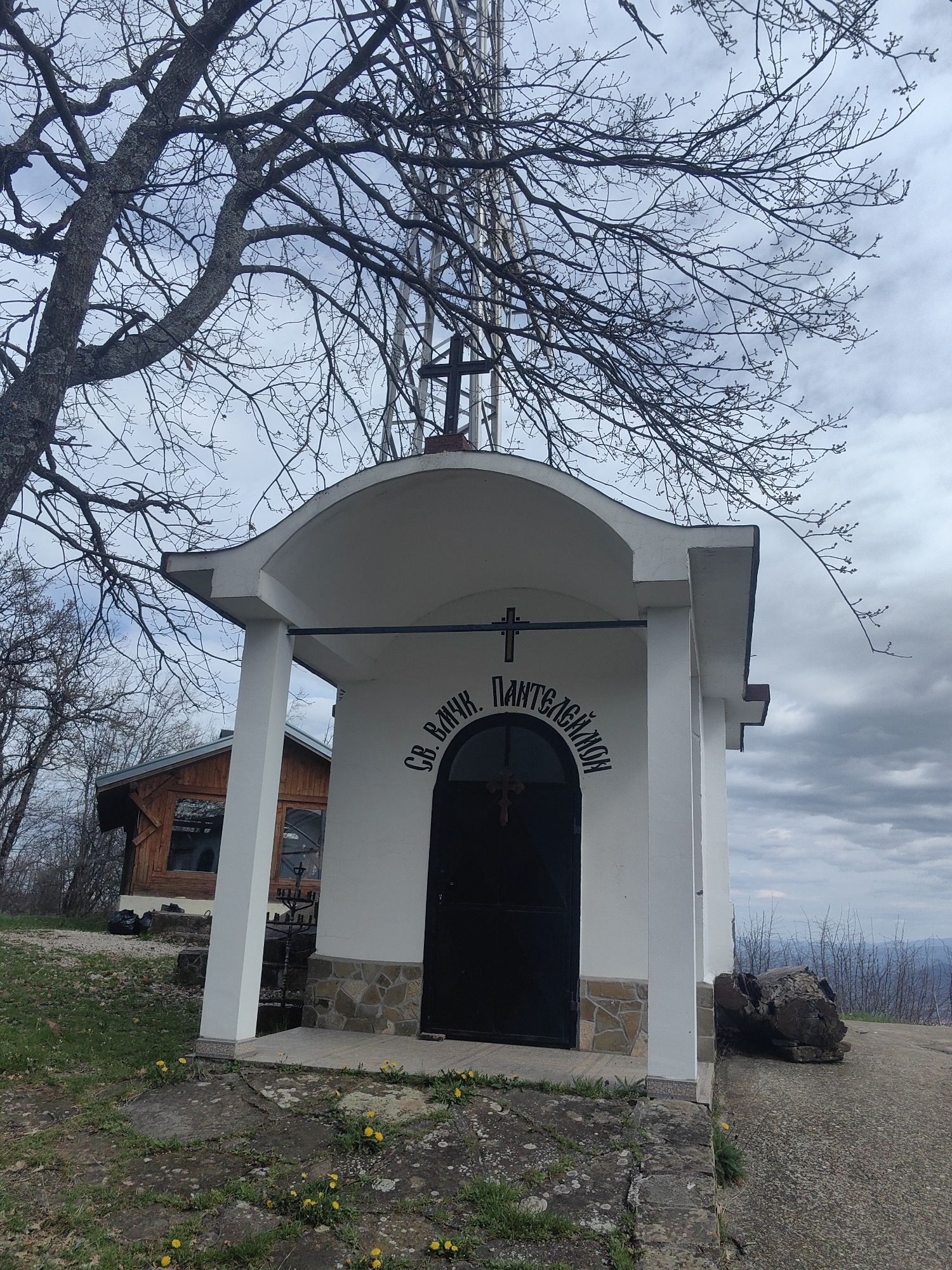 The image shows a small, white, rectangular Orthodox chapel with a dark arched door. Above the door, Cyrillic text reads "Св. Вмчк. Пантелеймон". A cross sits atop the roof. The chapel has two white pillars supporting a portico. The base of the chapel is decorated with stone cladding. The surrounding ground is stone and grass with a few yellow dandelions. Behind the chapel to the left is a small wooden cabin. In the background, there is a landscape of rolling hills under a cloudy sky. Bare trees surround the chapel. A metal tower is visible partially obstructed by a large tree. This building is located in Gumoshtnik, Bulgaria.