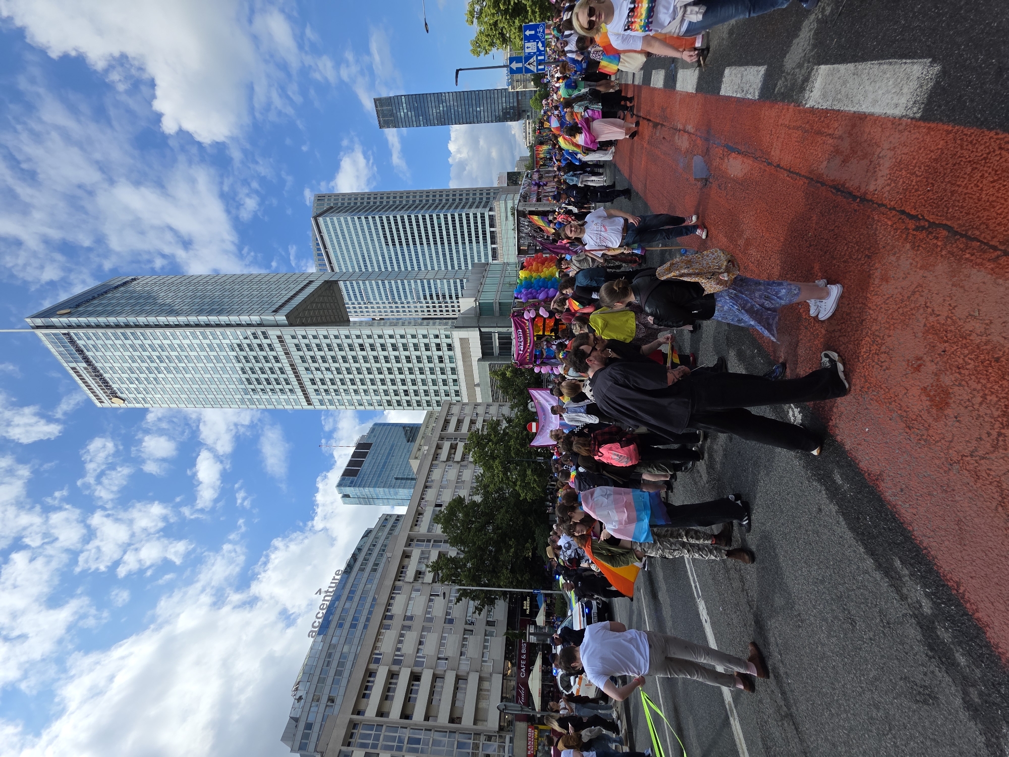The image shows a wide street in Warsaw, Poland, during a pride parade. In the background, a very tall, modern skyscraper dominates the skyline. To the left of the skyscraper are several other buildings, including one with the "accenture" logo visible. The street is filled with a crowd of people, many of whom are holding flags and banners. Several people are wearing colorful clothing, and rainbow flags are prominently displayed. Some individuals are holding what appear to be transgender flags. A police car is visible in the middle ground, along with a police officer. The road has a distinct red painted section and white crosswalk markings. The sky is blue with scattered white clouds. The perspective is from street level, looking upwards towards the buildings and the crowd.