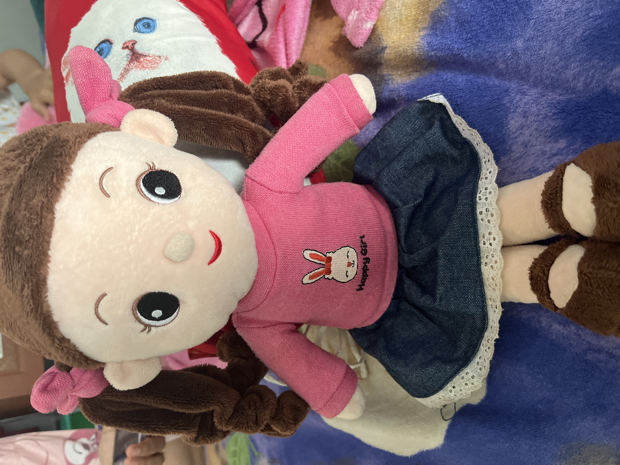 A stuffed doll with brown braided pigtails tied with pink bows sits on a blue and purple fuzzy blanket. The doll wears a pink long-sleeved shirt with a white bunny embroidered on the front, which says "Happy Girl" below it. The doll also wears a denim skirt with a white lace trim. The doll has large, dark eyes and a red smile. Its feet are covered in brown fuzzy shoes. In the background, there is a red pillow with a white cat face on it and a pink fuzzy blanket. The image is taken at a close-up, eye-level perspective.