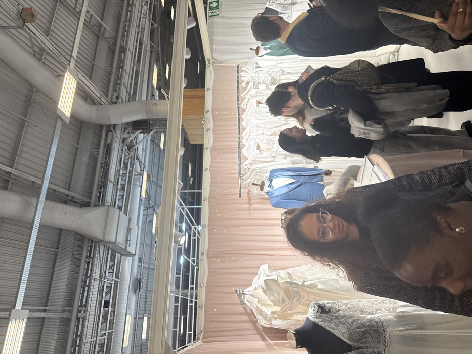 The image captures an indoor scene, likely at a wedding fair or bridal expo, with people browsing wedding attire. The overall image is rotated 90 degrees clockwise.
To the left of the image, a large industrial-style ceiling is visible, characterized by exposed white and grey ductwork, pipes, metal support beams, and bright rectangular fluorescent lighting fixtures. A small cardboard box is attached to one of the ceiling structures in the upper left. A green fire exit sign with a white running person symbol is also visible against this industrial background.
To the right, a booth or display area is set up against a soft, light pink backdrop, possibly fabric drapes, which appears to have subtle sparkling details, like fairy lights or glitter. Within this booth area, several wedding dresses are displayed on mannequins or hangers. These dresses are predominantly white or off-white, with some featuring intricate lace details. A light blue suit jacket with a bow tie is prominently displayed on a mannequin in the middle ground, and further back, a green suit jacket is also visible on another mannequin.
Several people are present in the booth area, engaged in browsing the displayed items. In the immediate foreground, a woman with long, dark, curly hair and glasses is looking towards the viewer. She is wearing a dark, possibly patterned, jacket. Partially visible to her right is another person with shorter, dark hair and an earring, facing away from the camera.
Further back in the booth, several other individuals are observed. One person is seen from behind, with dark, curly hair, wearing a dark jacket over a lighter shirt, holding what appears to be a brochure or pamphlet. Next to them, another individual is seen from a side/back angle, wearing a dark jacket and carrying a patterned tan and dark shoulder bag. They seem to be looking at the wedding dresses. Behind these individuals, another person with long, dark hair is also visible, observing the displays. In the far back, an individual with short, dark hair, wearing a dark blue top with a light-colored collar, is seen from behind, looking at the green suit jacket display.
The lighting in the booth area appears softer and warmer than the stark fluorescent lighting of the industrial ceiling area, creating a distinct separation between the exhibition space and the venue's structure. The overall activity suggests a casual browsing experience at an event focused on wedding planning or apparel.