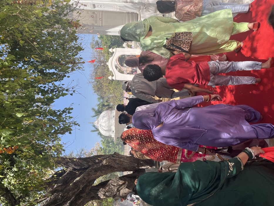 The image shows a group of people walking on a red carpet outside a white building in Panchkula, India. The building appears to be a religious structure, with a white dome and an archway entrance. The people are dressed in traditional Indian clothing, and some are carrying bags or purses. There are trees to the left and right of the building. The image was taken at the coordinates 30.721 latitude, 76.854 longitude. The scene is outdoors in daylight.