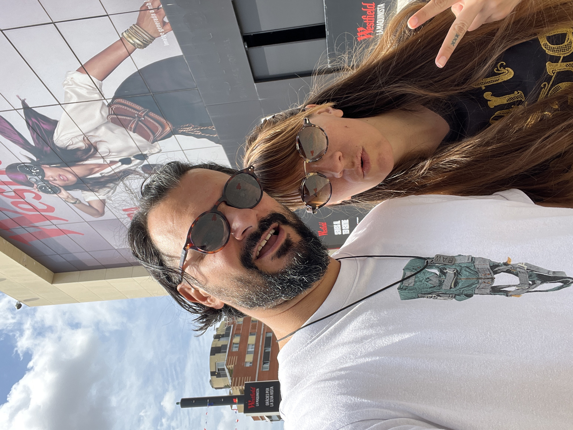 A selfie taken outdoors in front of a large billboard for Westfield La Maquinista. A man with dark hair, a beard, and sunglasses is in the foreground on the left, wearing a white t-shirt with a graphic print and a necklace. Next to him on the right is a woman with long brown hair and glasses, wearing a dark t-shirt and making a peace sign with her right hand, which has a tattoo on it. The billboard features a fashion advertisement. The sky is visible with clouds, and there are buildings in the background, consistent with an urban environment in Barcelona, Spain.
