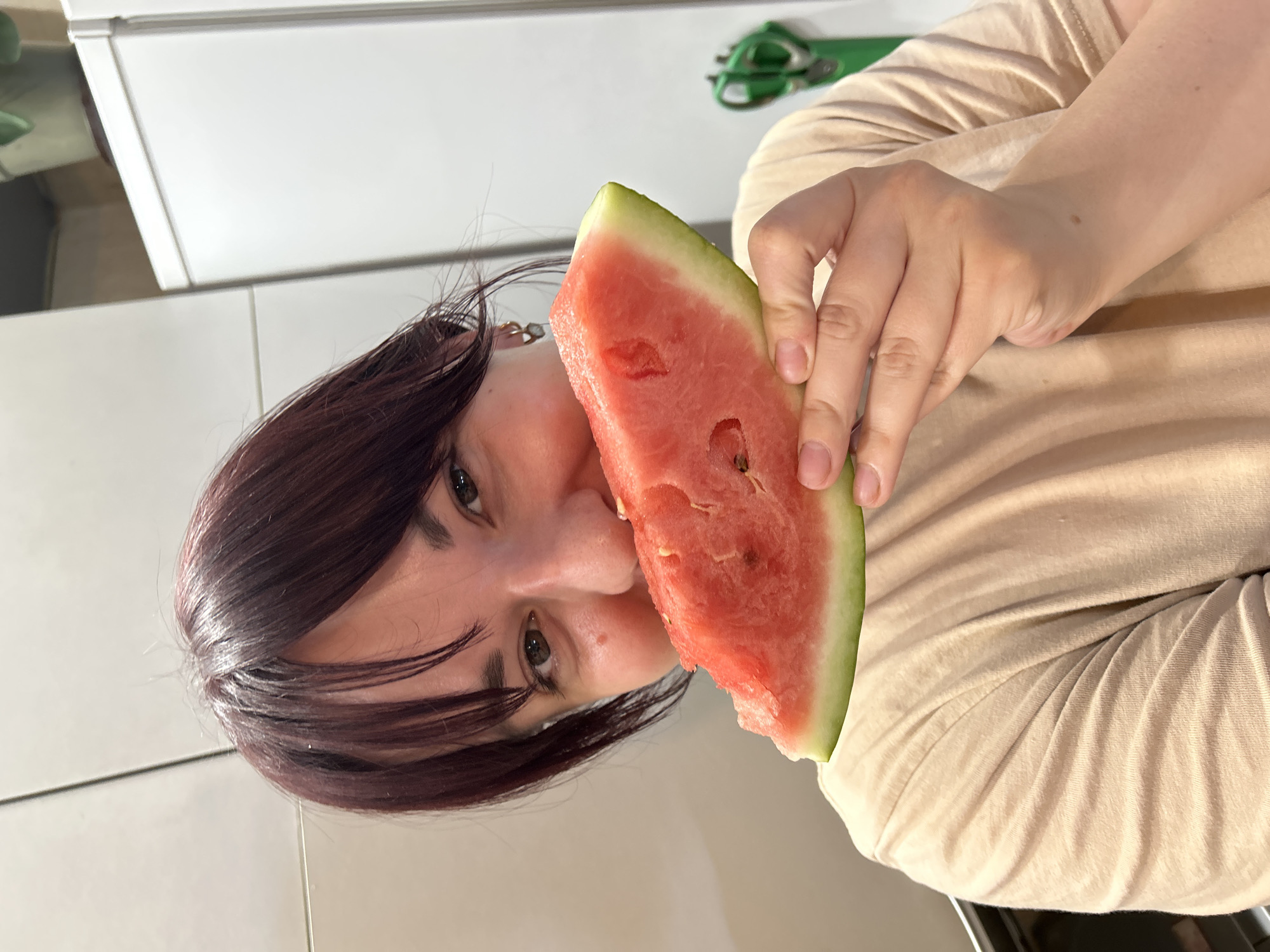 The image shows a woman with dark hair holding a slice of watermelon to her face. She is wearing a beige t-shirt. The background appears to be a kitchen, with a white refrigerator and cabinets visible. There is also a pair of green scissors on a shelf in the background. The woman's face is partially obscured by the watermelon. The lighting suggests an indoor setting. There is no visible indication of the location being in the city of Фоки, Russia.