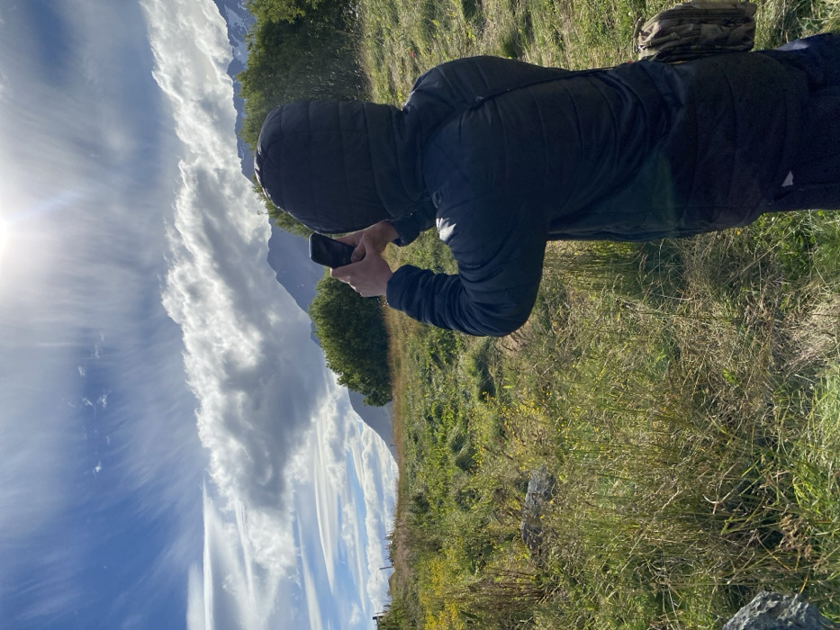 A person wearing a black hooded jacket takes a picture with their phone of a landscape. The sky features wispy clouds and a bright sun on the left. Mountains and trees populate the background. A grassy field with patches of yellow flowers makes up the foreground.