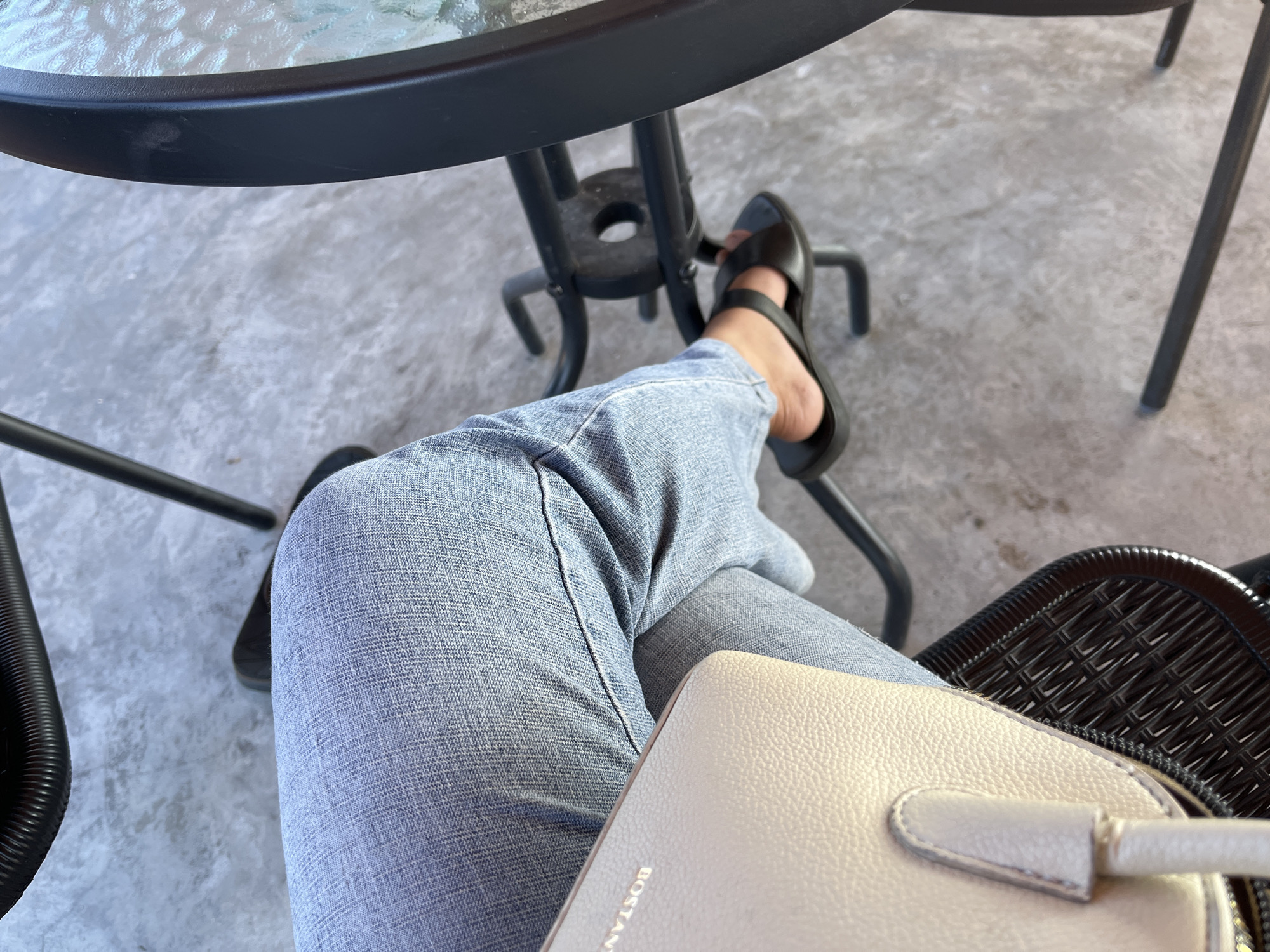 This is a high-angle, eye-level, medium shot of a person's legs and a portion of a purse, placed in front of a table and chair. The person is wearing light blue denim jeans and black slide sandals. The purse is beige with a textured surface and the word "BOSTANT" is embossed on it. The background consists of a concrete-like surface with a woven black chair and a dark-colored table with a glass top. The scene appears to be outdoors. No specific location in Oro-oro Ombo, Indonesia is identifiable.